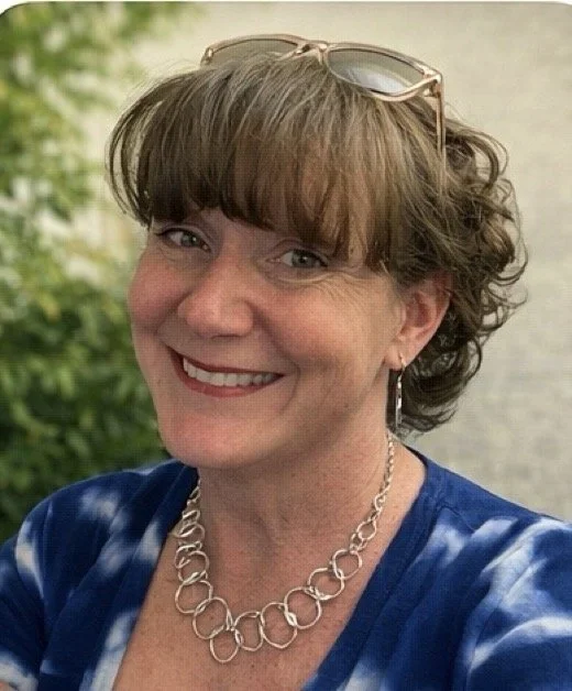 A smiling woman with short, curly hair, wearing glasses on her head, a blue tie-dye shirt, a silver chain necklace, and earrings, standing outdoors with greenery and a body of water in the background.