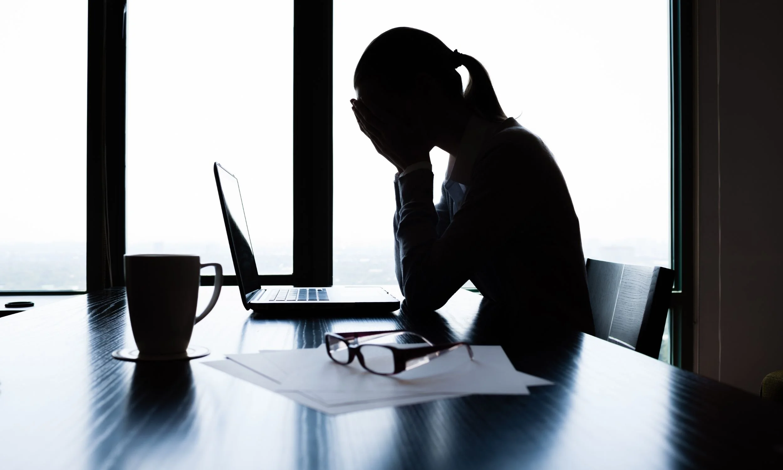 Persona con duda en un escritorio con una computadora portátil, gafas y documentos, en un ambiente con ventanas grandes y luz natural.