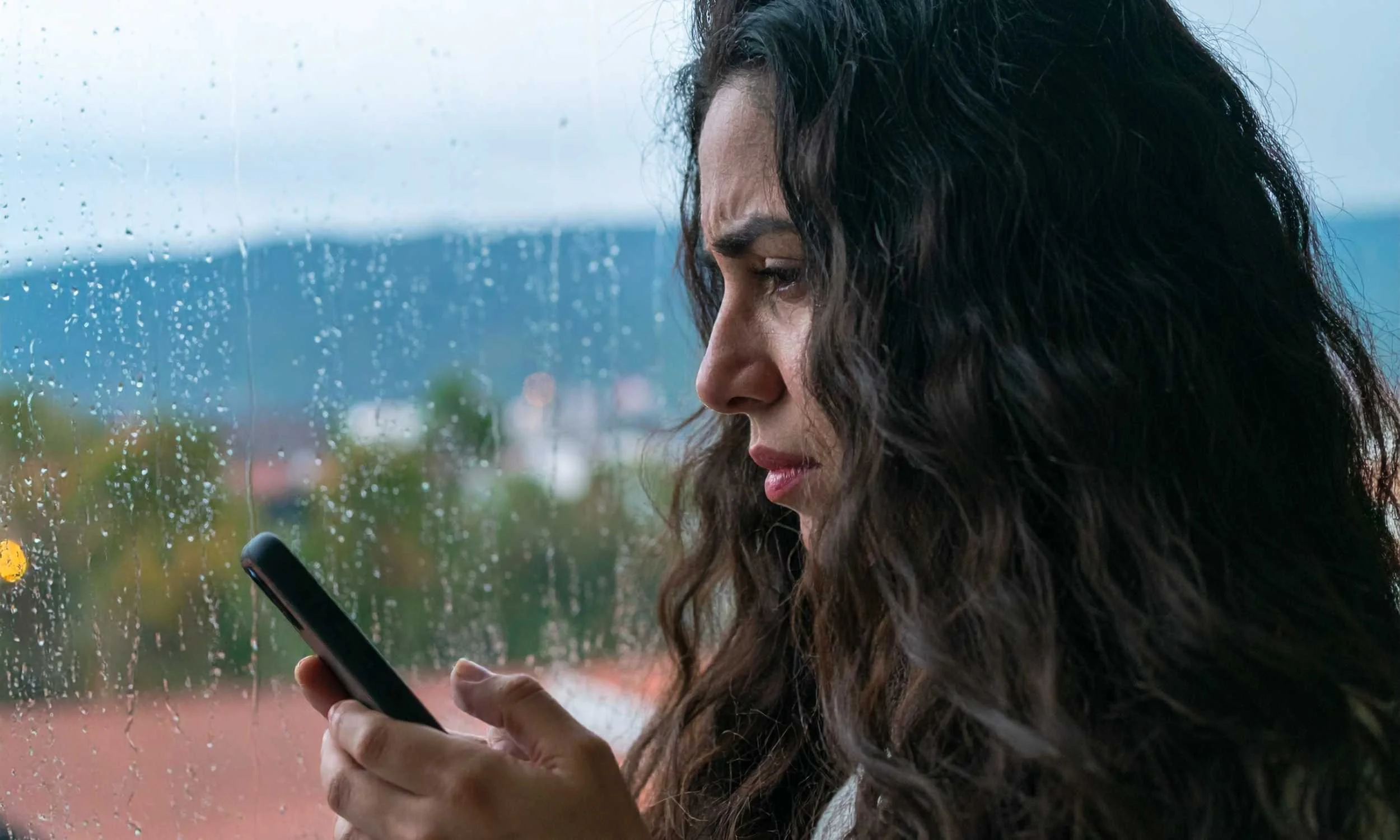 Mujer con cabello rizado mirando su teléfono móvil en un día lluvioso, vista a través de una ventana con gotas de agua.
