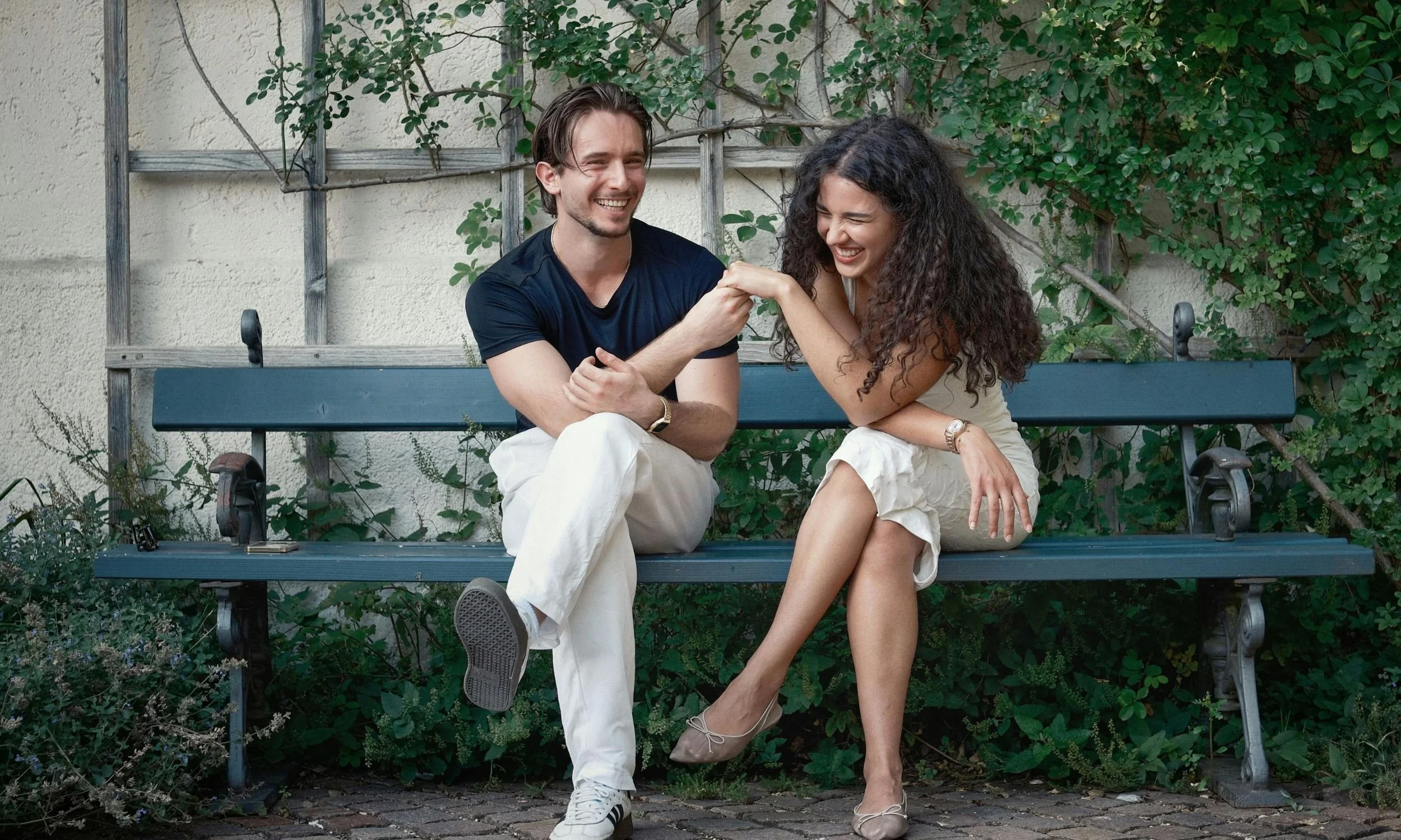 Una pareja joven riendo y jugando en una banca de parque, tocándose las manos en señal de amistad o competencia, rodeados de vegetación.