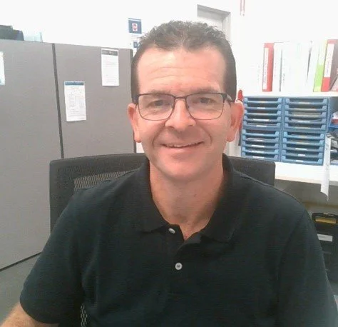 A man with short dark hair and glasses sitting at a desk in an office environment.