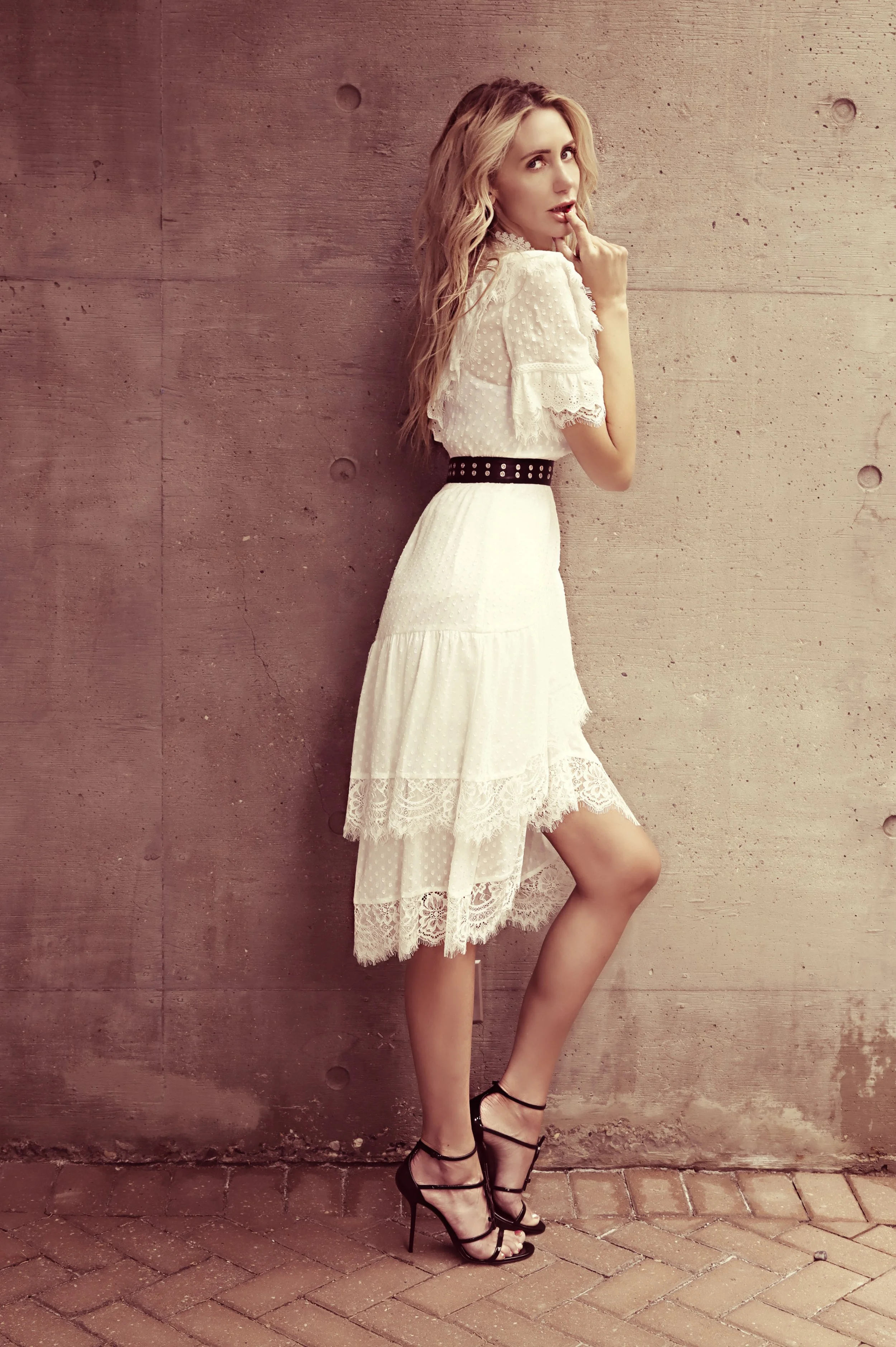 A woman in a white lace dress with a black belt and black high heels standing against a concrete wall. She is looking over her shoulder with one finger near her lips.