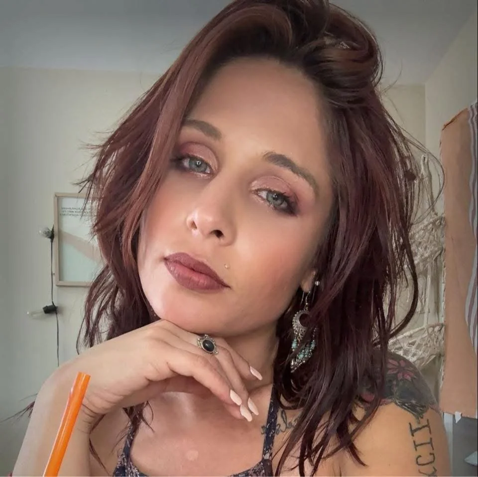 A woman with reddish-brown hair, blue eyes, and hoop earrings, wearing a black lace top with thin straps.