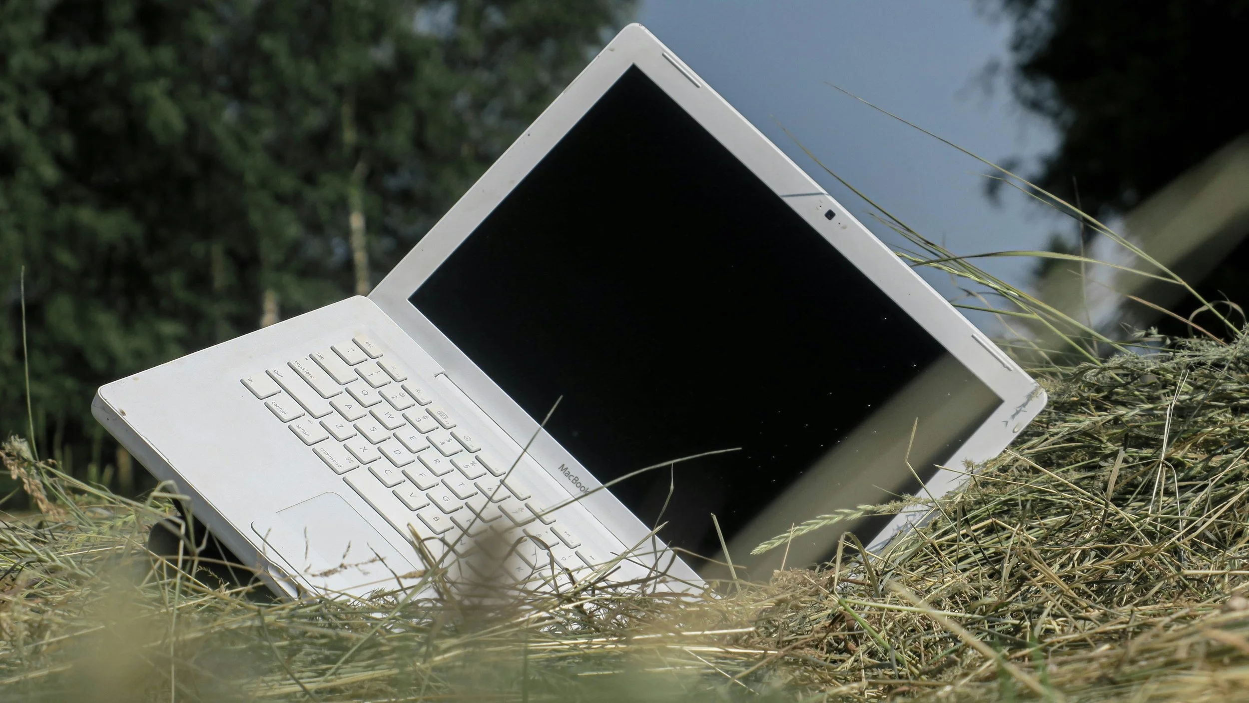 White laptop with black screen and keyboard resting on dry grass outdoors, with trees and water in the background.