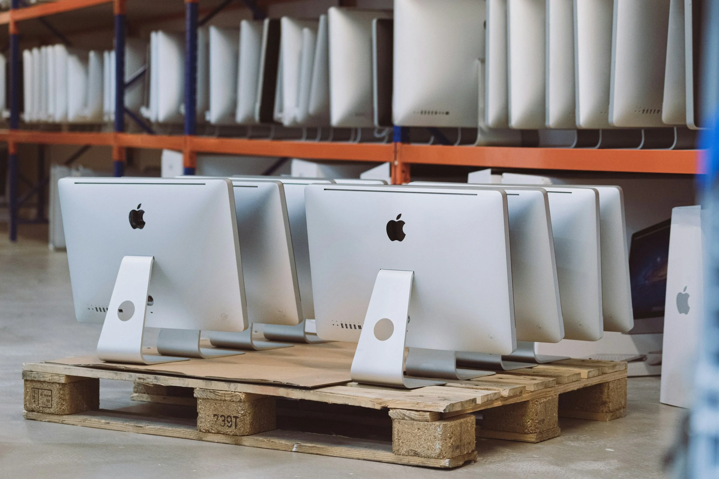 Several white Apple iMac computers are arranged on a wooden pallet in a warehouse, with shelves of more computers visible in the background.