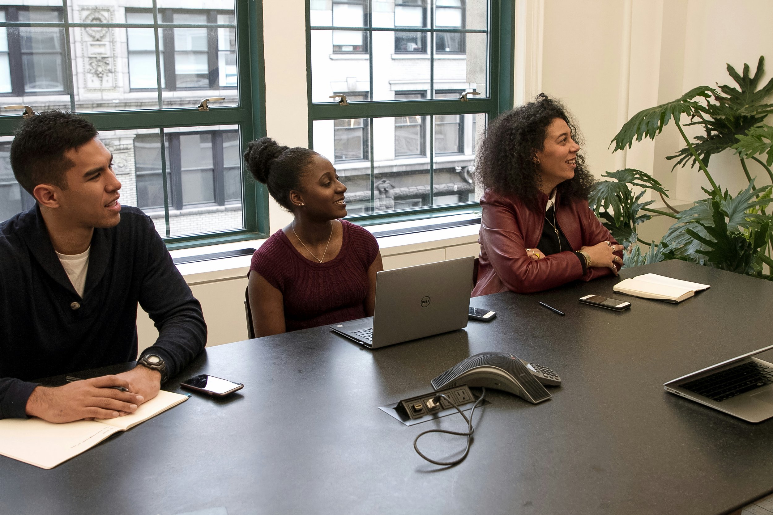Three diverse individuals sitting at a conference table in a meeting room, with laptops, notebooks, and phones in front of them, engaged in discussion.