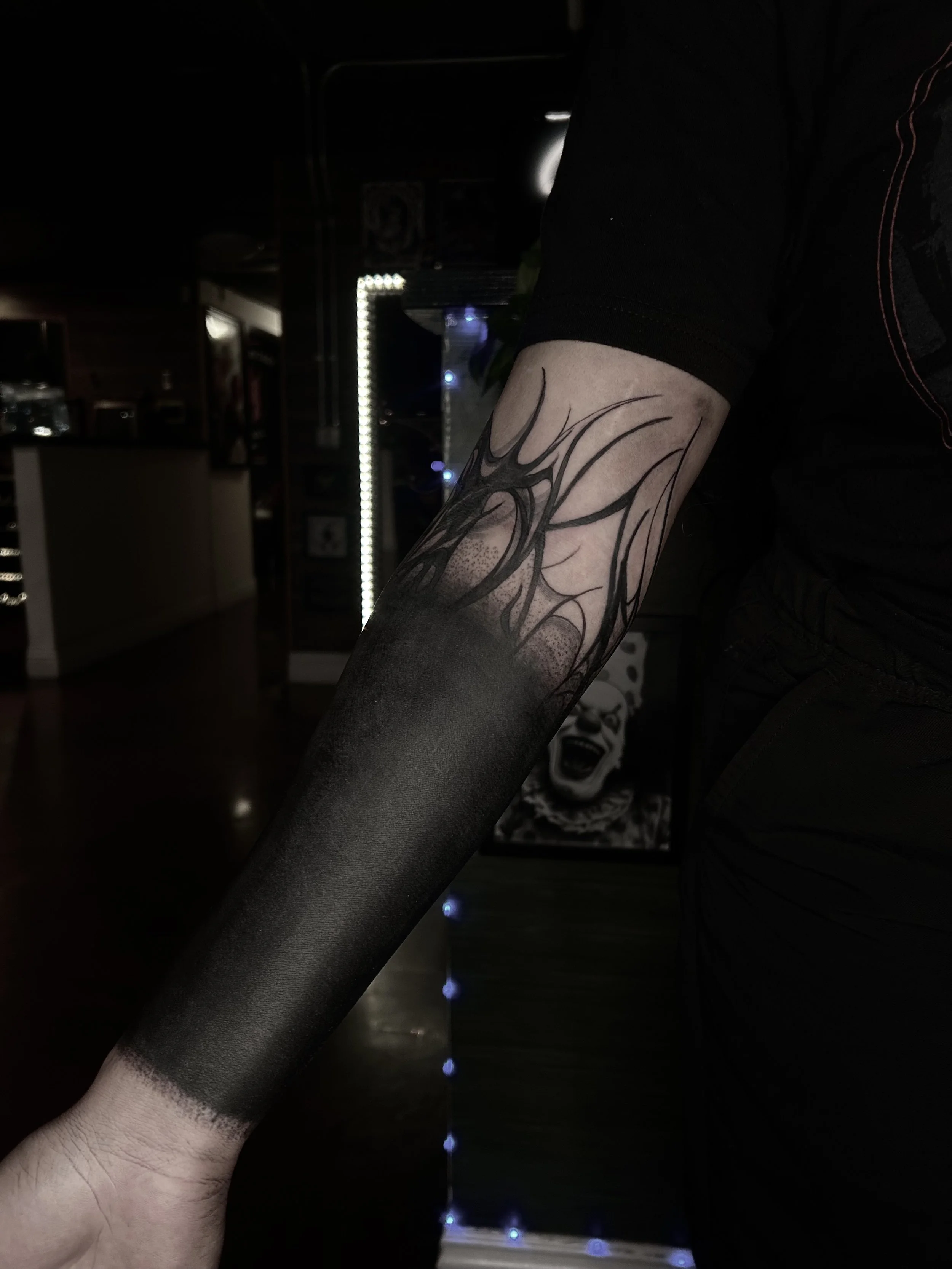 Tattooed arm with black and grey shading and abstract flame-like design in a dimly lit indoor setting.