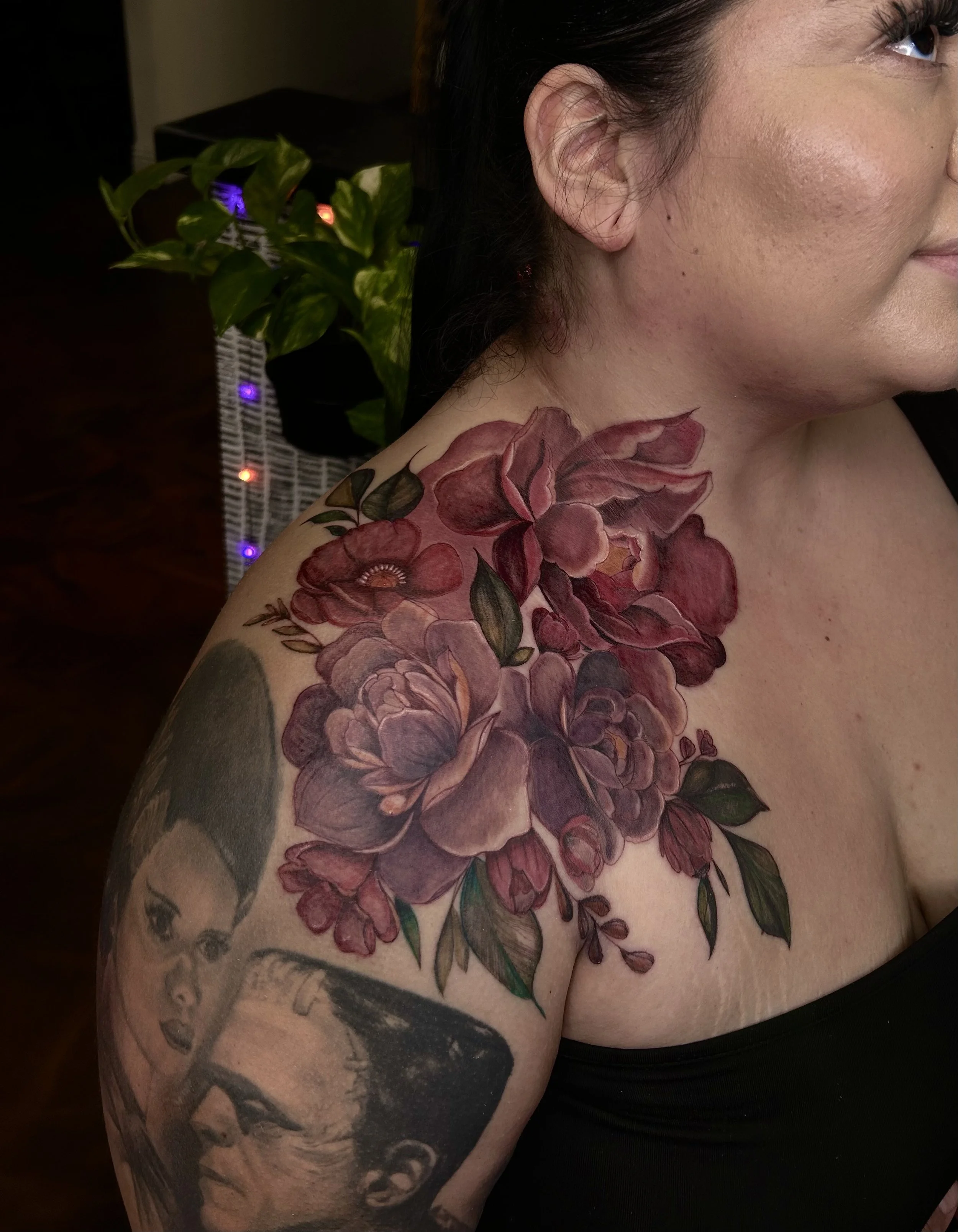 A woman with a floral tattoo on her left shoulder and upper arm, featuring large pink and purple flowers with green leaves, and black and white portrait tattoos of a woman and a man on her arm.