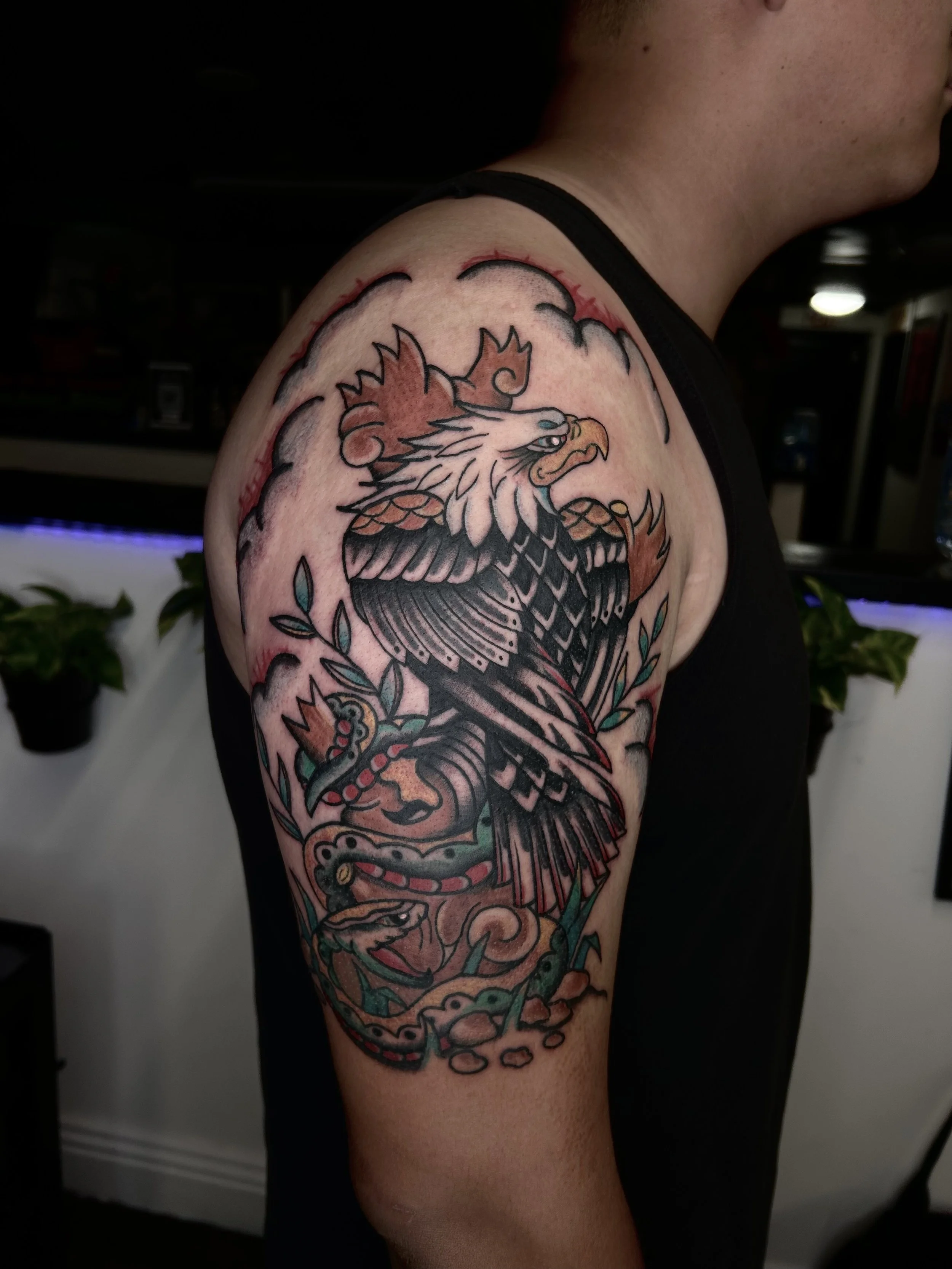 Tattoo of an eagle with wings spread, holding a snake in its talon, surrounded by clouds and leaves.