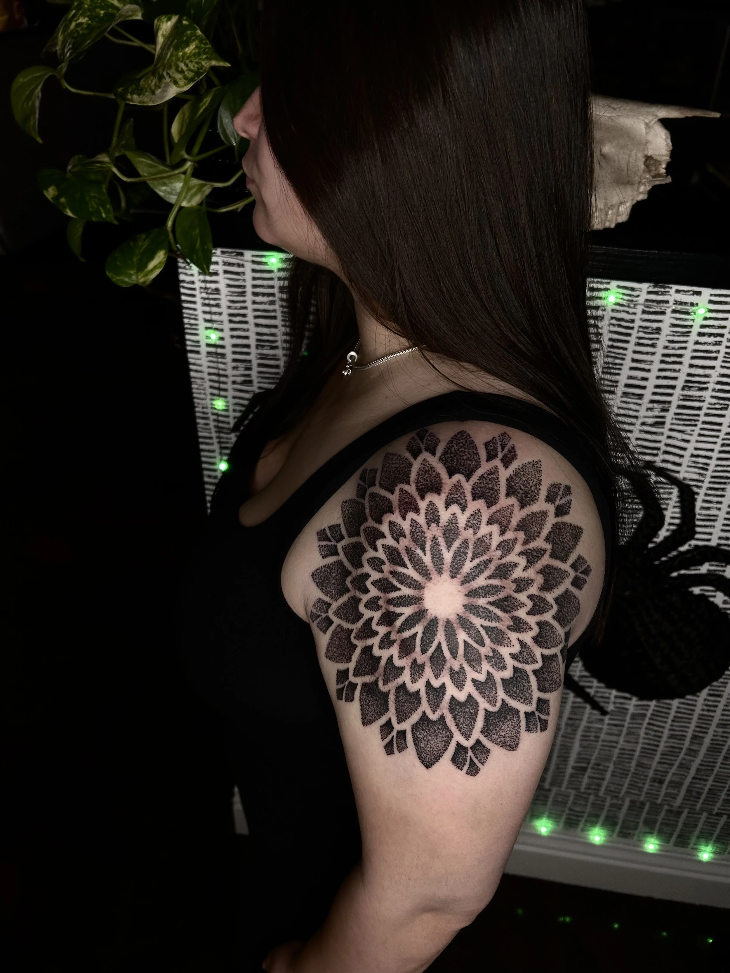 A woman with long dark hair showing a large black and pink floral tattoo on her shoulder and upper arm, standing in front of a white wicker surface with green LED lights, and a potted plant in the background.