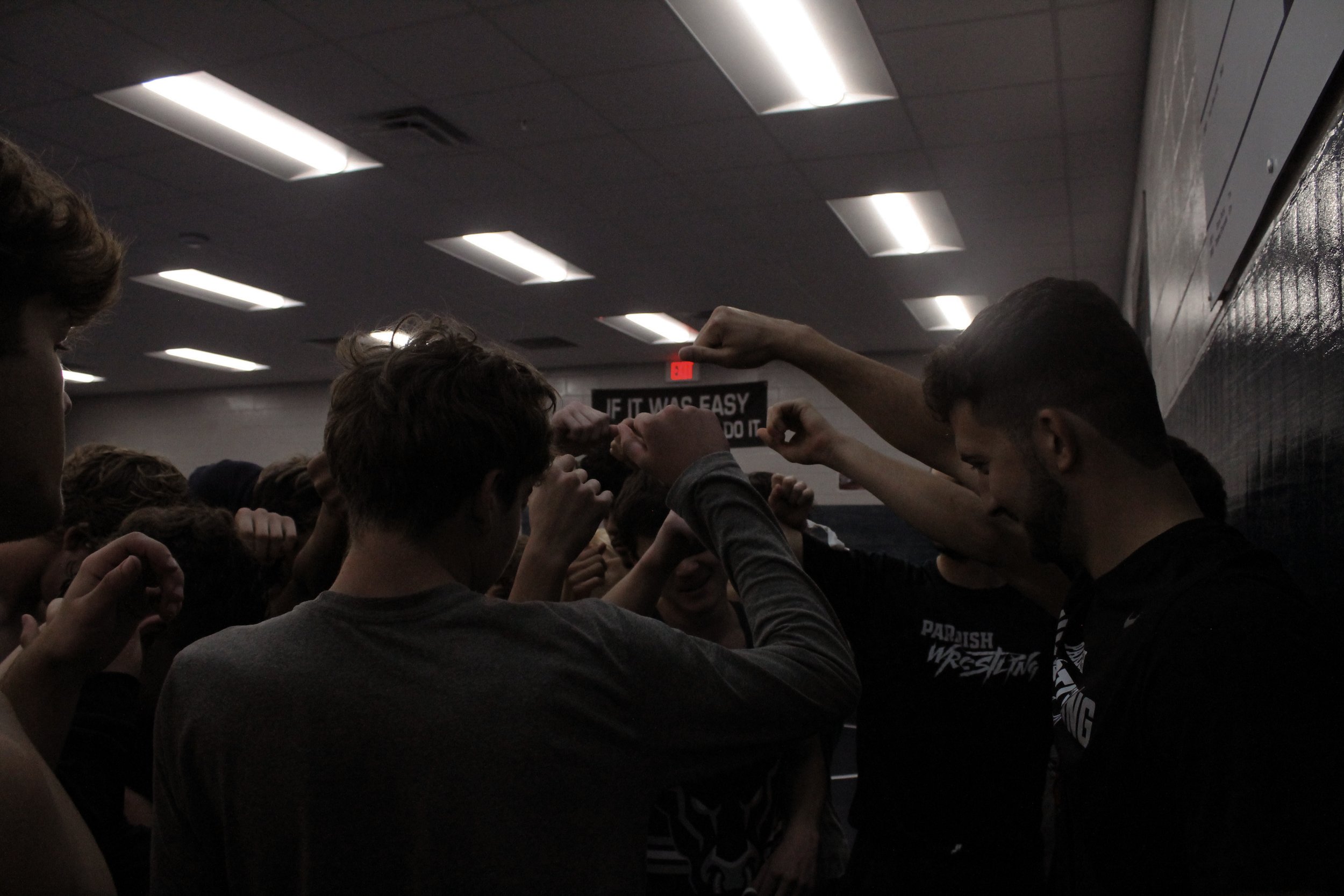 A group of people, likely athletes, gathered in a gym or locker room, raising their fists together in a team huddle.