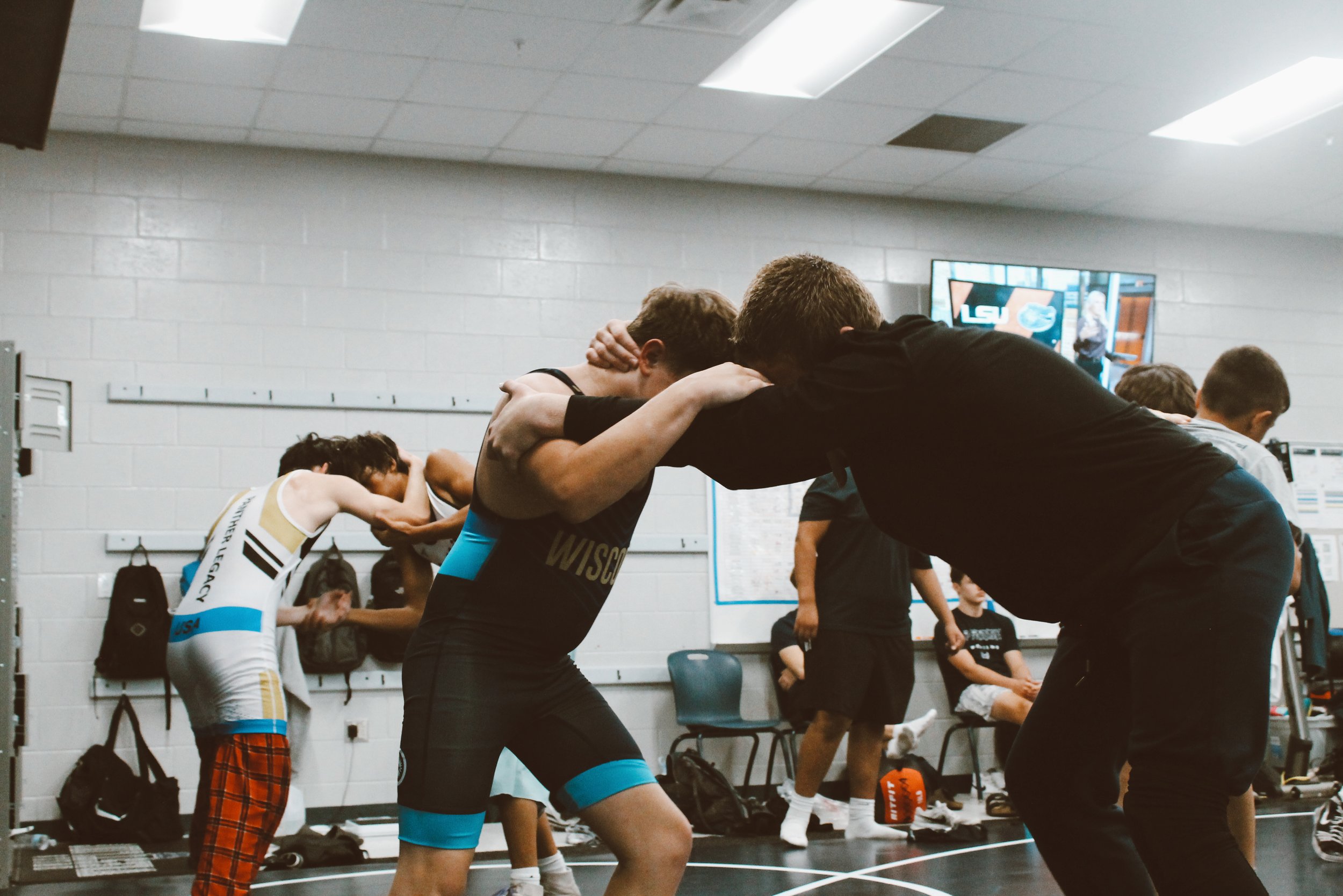 Wrestling practice with two young male wrestlers practicing moves, one in black singlet and the other in blue and black singlet, in a locker room with other young people and coaches in the background.