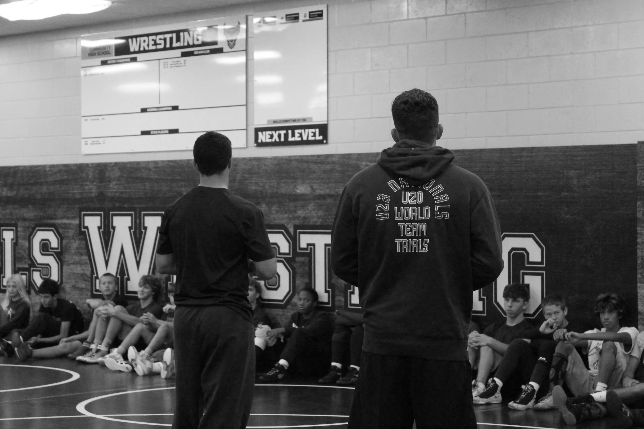 Two coaches standing in front of a group of young athletes seated on the floor of a gym. The gym wall has large lettering and a scoreboard. The scene is in black and white.