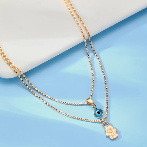 Embrace timeless protection and style with this Gold Plated Double Layer Evil Eye and Hamsa Hand Necklace from Mozzimo Jewelry.