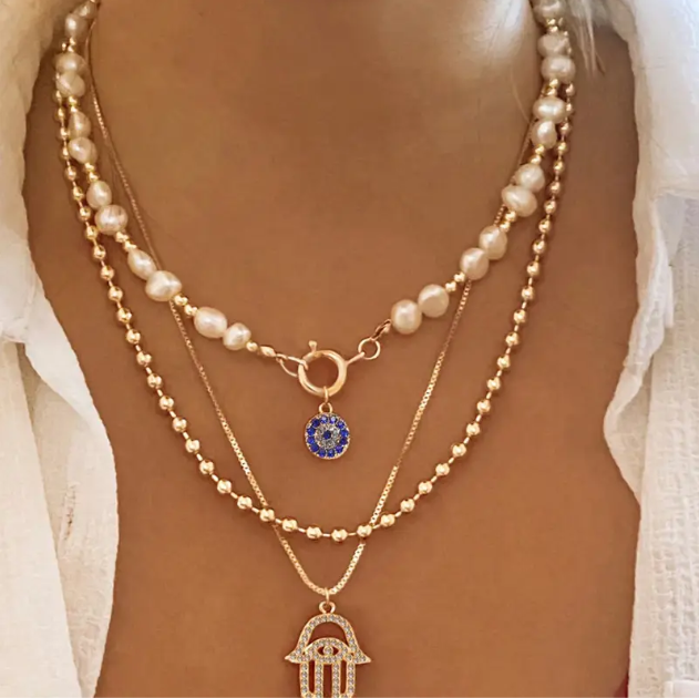 Elevate your look with this boho-chic gold plated triple layer necklace featuring pearls, beads, and a rhinestone Hamsa hand charm.