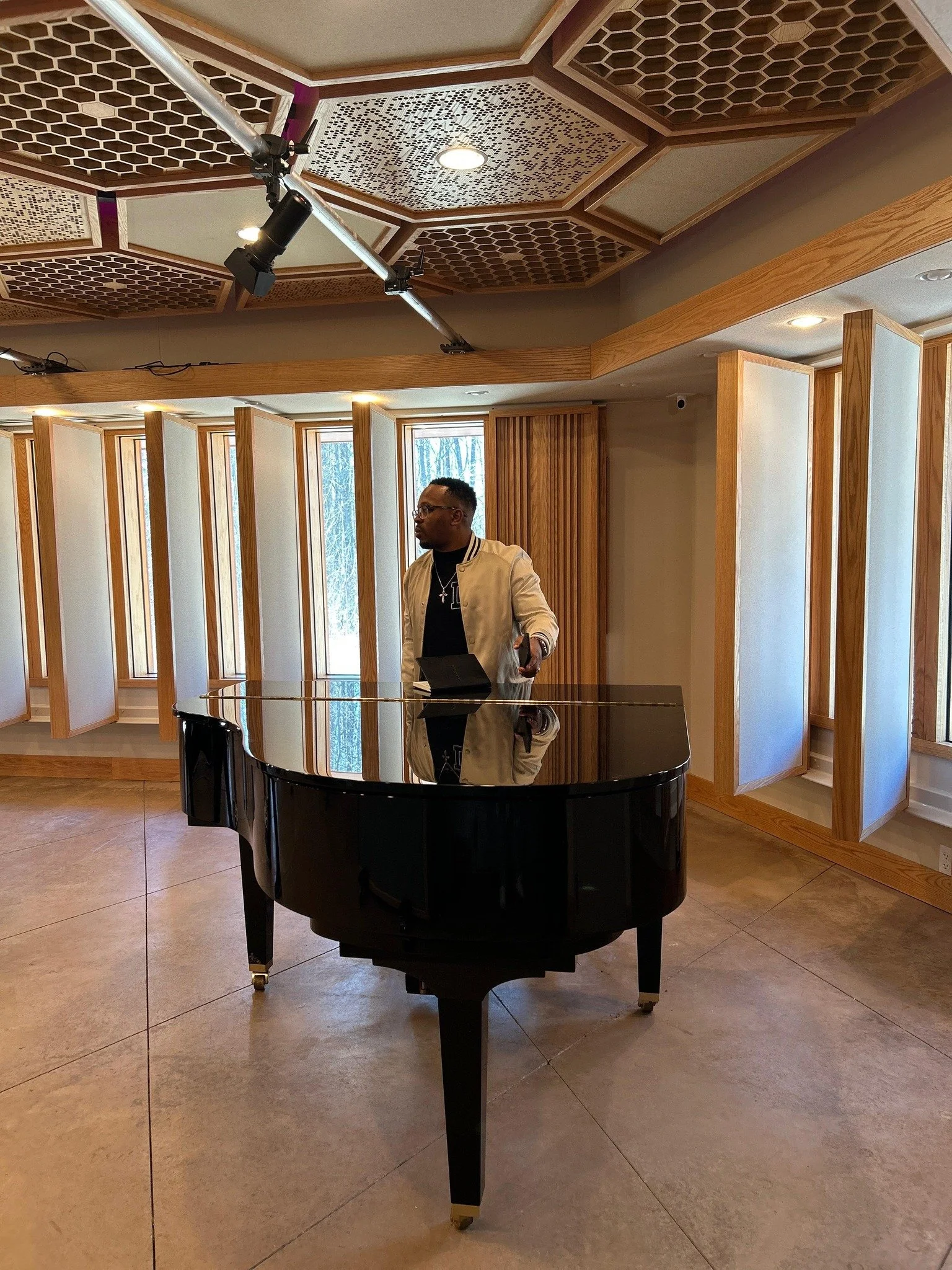 A man in a white jacket standing behind a black grand piano indoors, with a ceiling featuring acoustic panels and large windows with wooden frames.