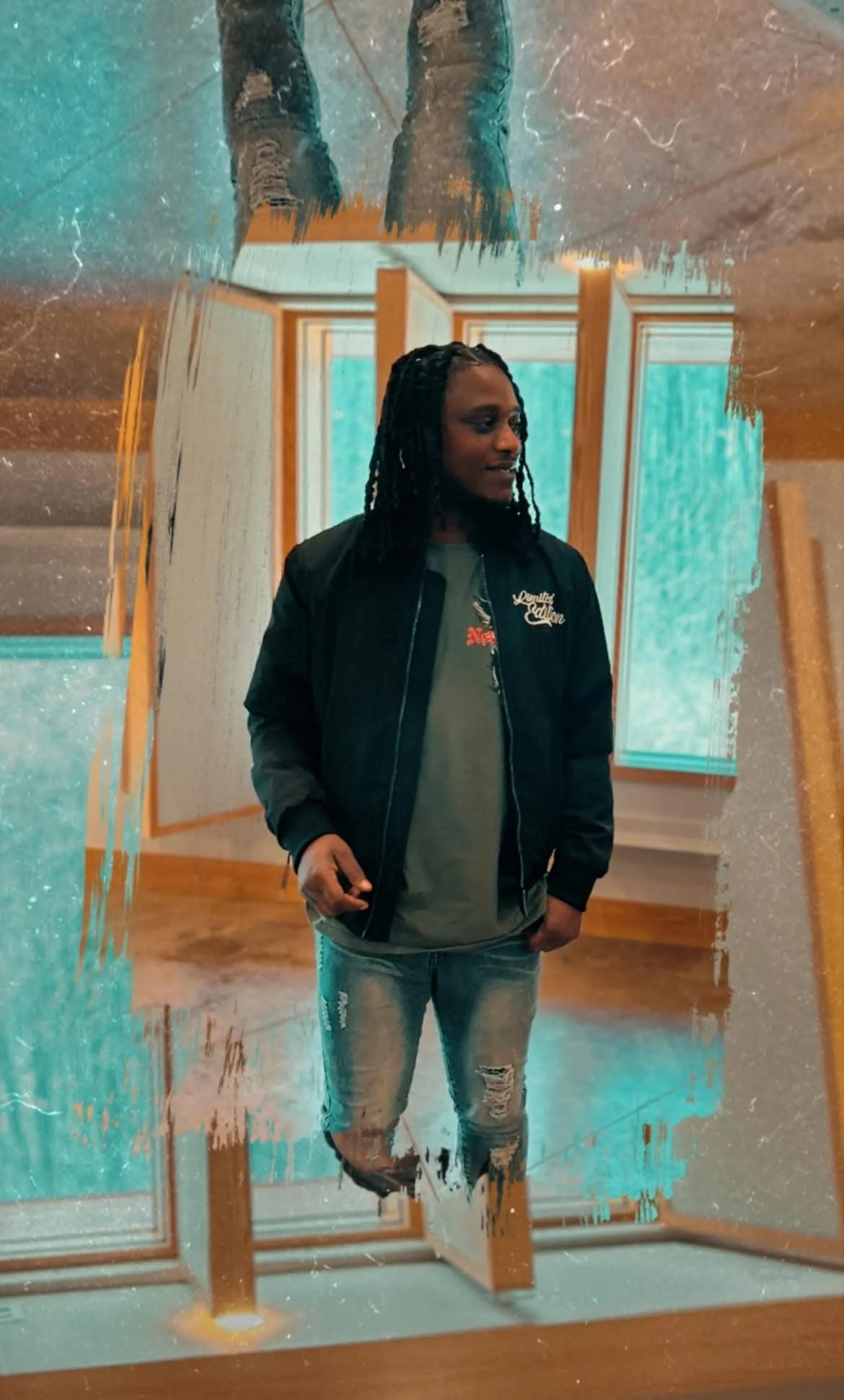 A person with long braided hair standing in front of a mirror, wearing a black jacket and ripped jeans, in a room with large windows and wooden floors.