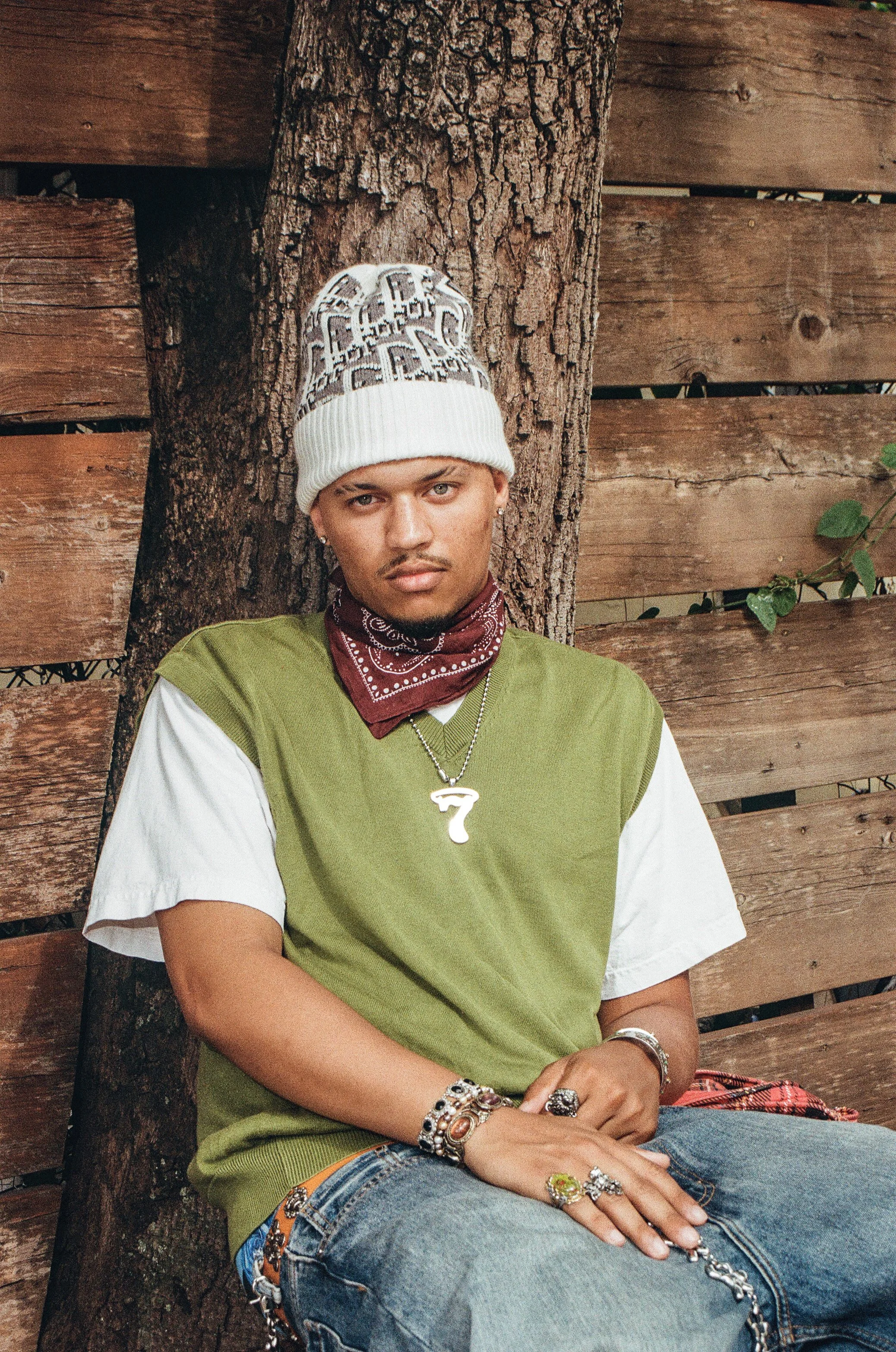 Young man sitting against a tree with a wooden fence in the background, wearing a white beanie, a maroon bandana, jewelry, a green and white t-shirt, and jeans.