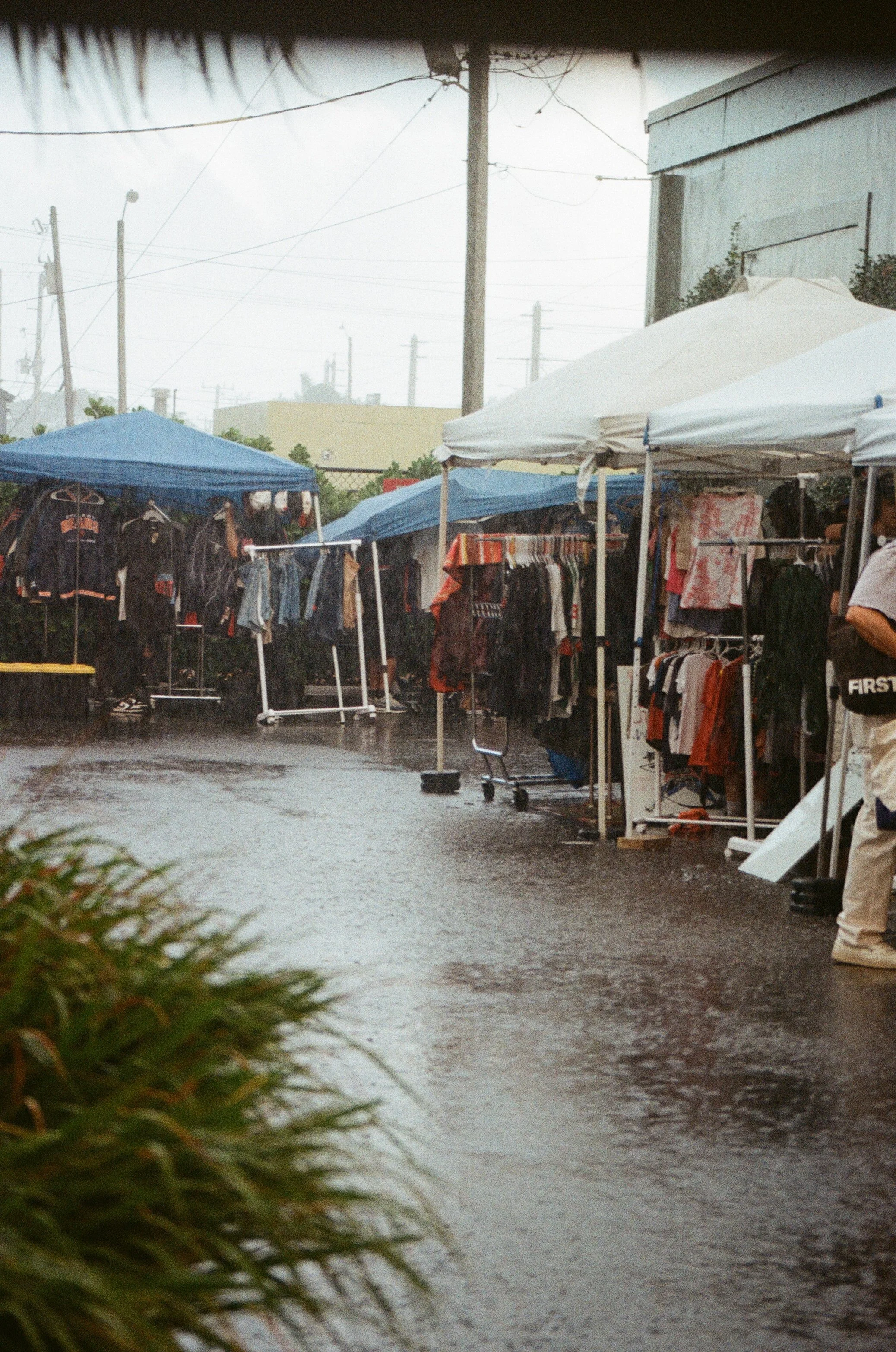 Outdoor clothing market with rain-soaked ground and tents, rain falling, various clothing items hanging on racks, and people browsing in rainy weather.