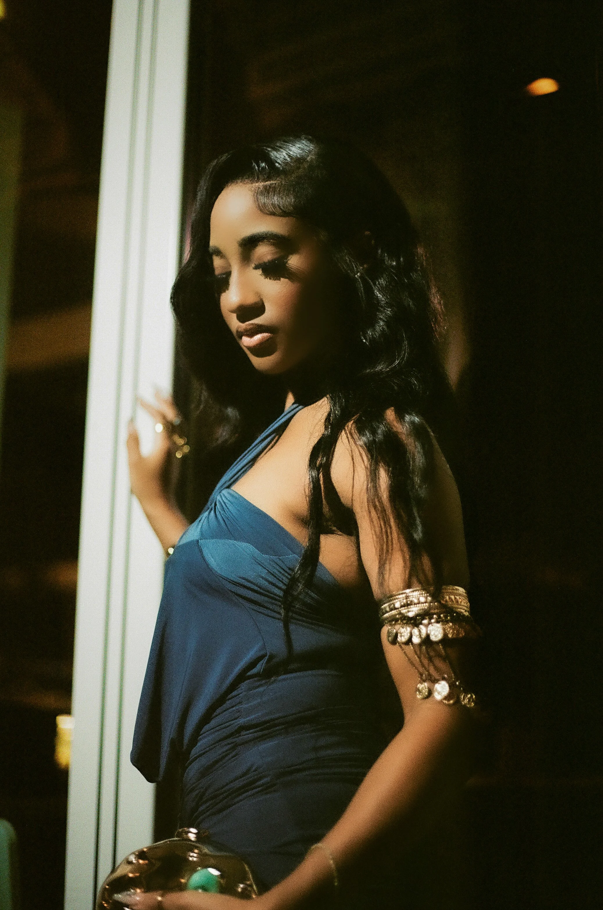 A woman with long black hair, wearing a dark blue dress and gold jewelry, poses indoors with closed eyes, leaning against a wall with a dark background.