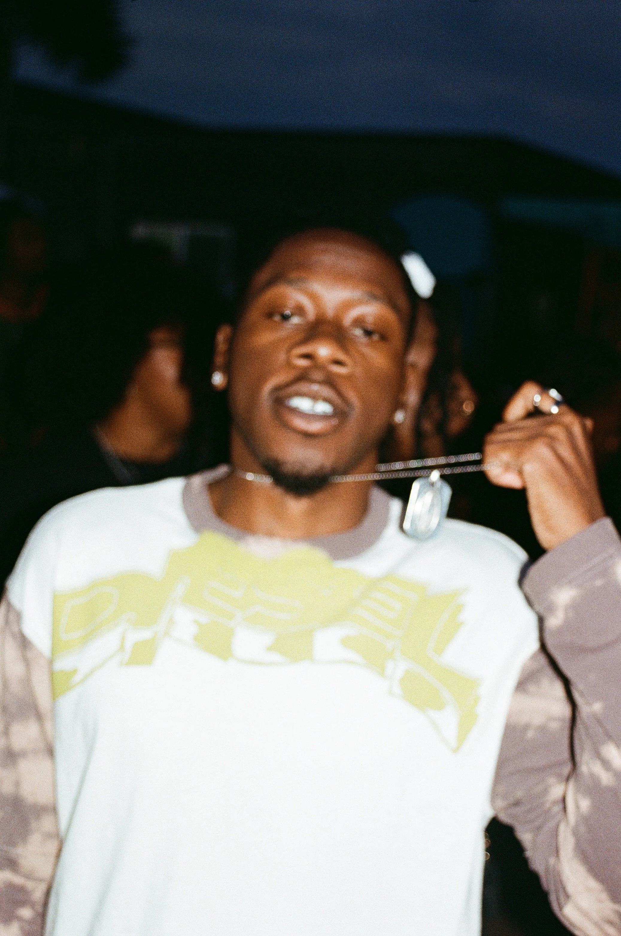 A young man with earrings and a chain necklace holding a dog tag chain near his neck, in a dimly lit setting with other people in the background, possibly at a party or club.