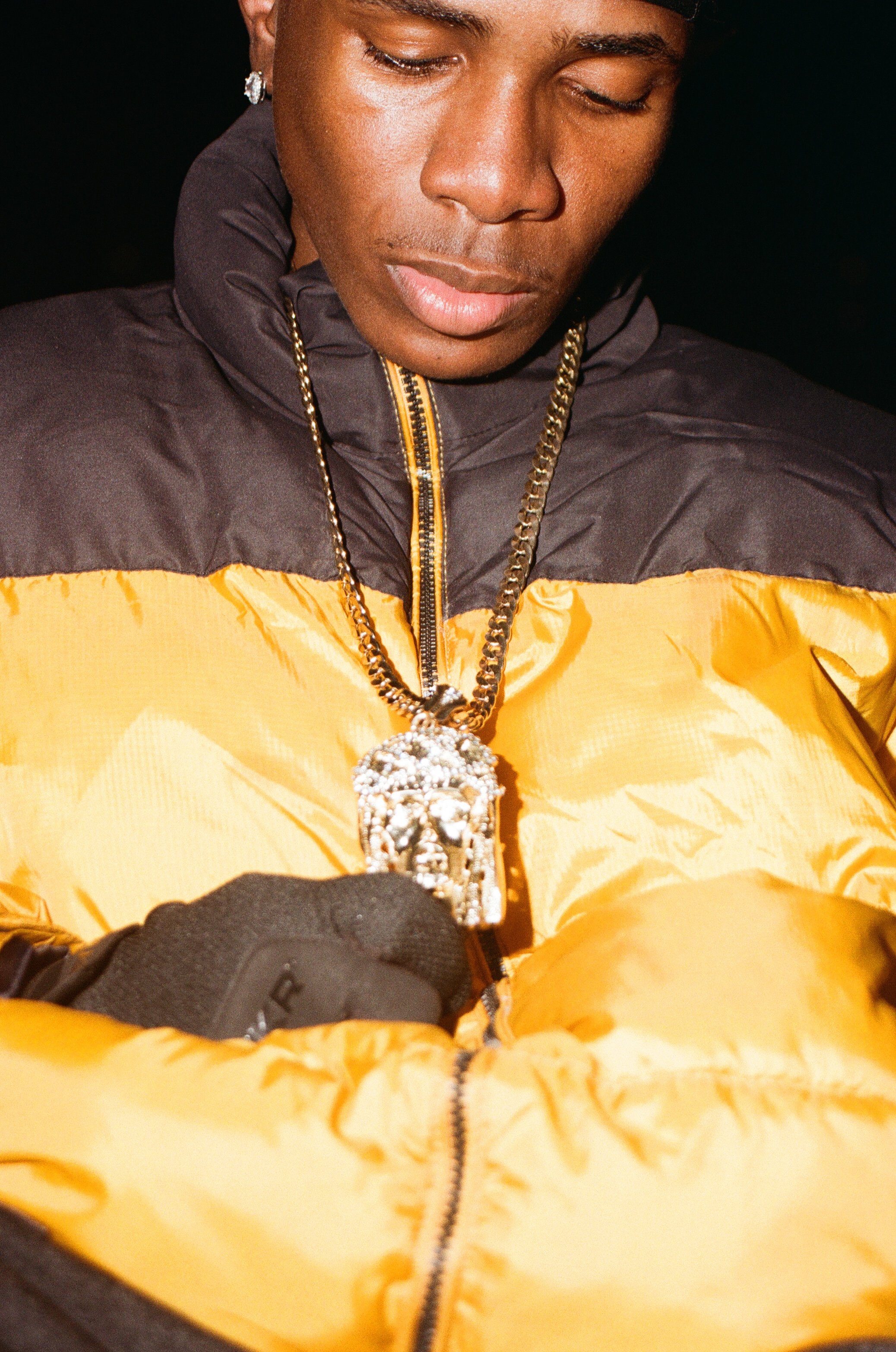 Close-up of a person wearing a yellow and black puffy jacket, diamond earrings, and a large diamond chain with a pendant, with their eyes closed and head slightly tilted downward.