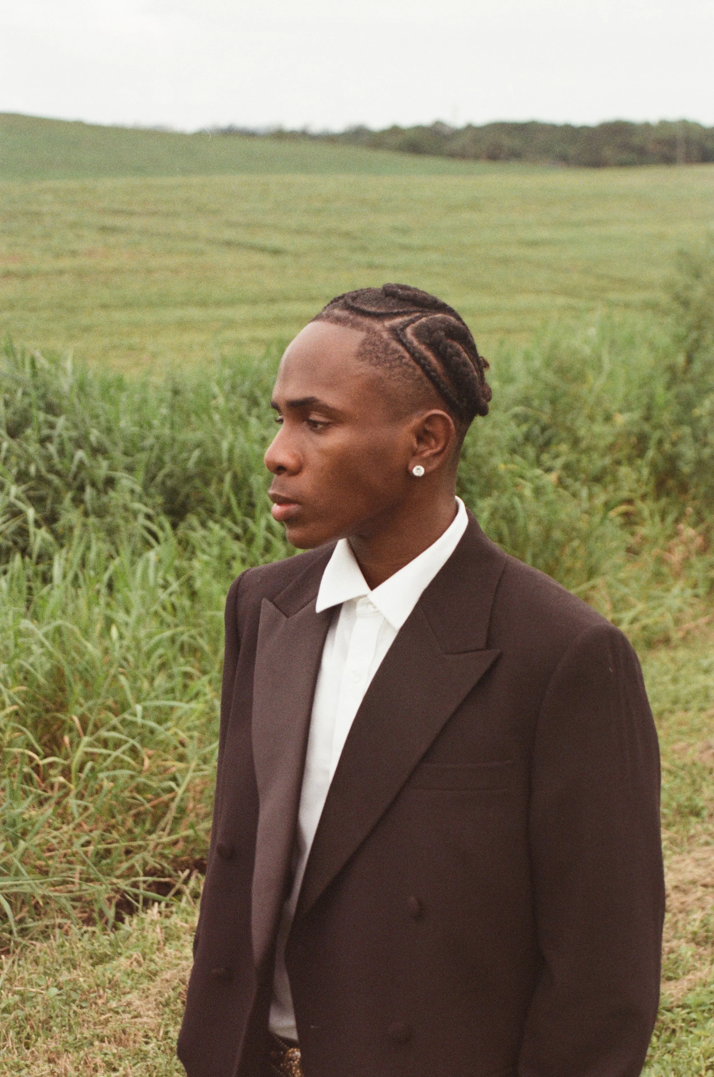 A young man in a black suit with a white shirt, standing outdoors in a green field with grass and shrubs, looking to the side with a serious expression, wearing earrings and styled hair.
