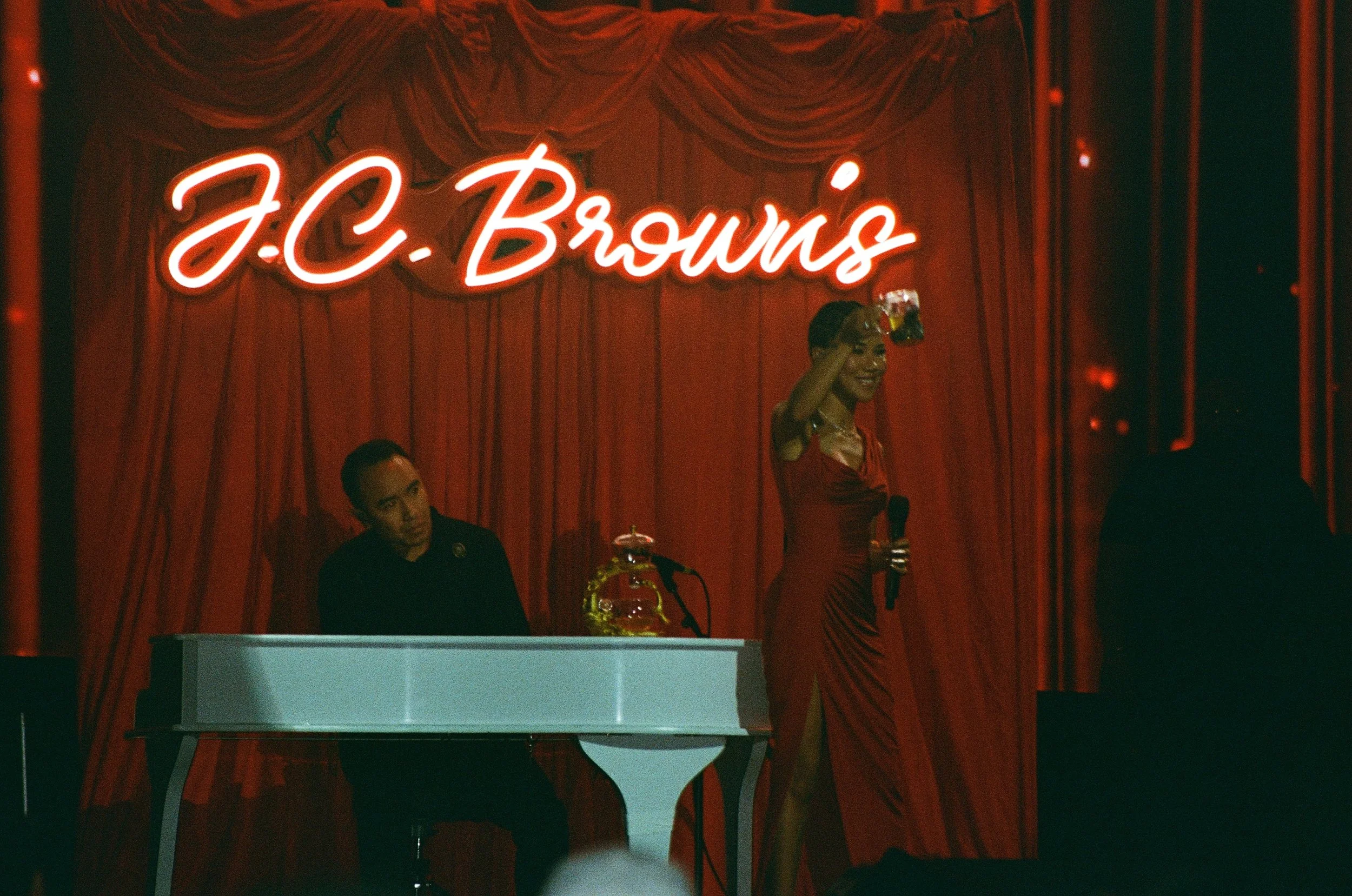 A woman in a red dress is holding a microphone and a glass of drink on stage, smiling, with a man sitting at a white piano behind her. The background features red curtains with a neon sign that reads 'J.C. Brown'.