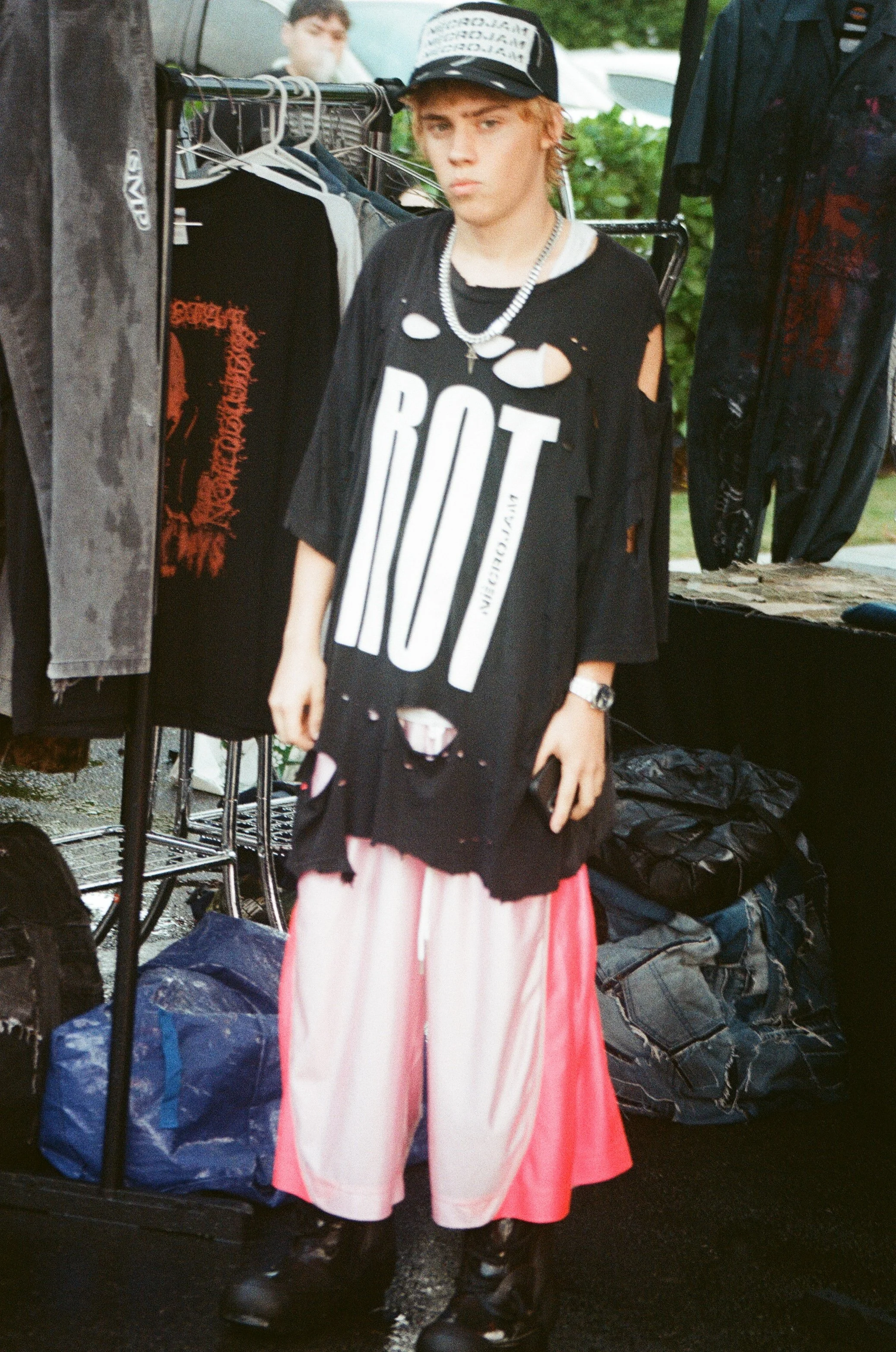 Teenager with bleached hair wearing a large black t-shirt with the word 'ROY' and ripped holes, pink and white wide-leg pants, black boots, and layered necklaces, standing among clothing racks at an outdoor market.