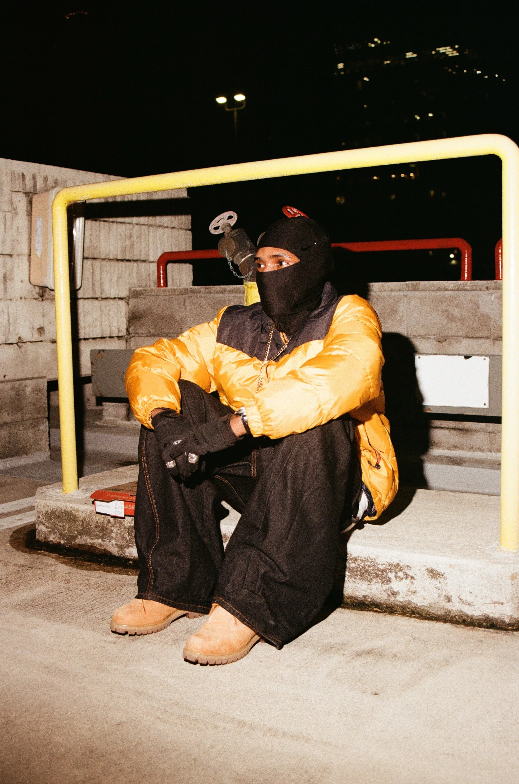 A person wearing a black face mask, black gloves, a yellow puffer jacket with black accents, black jeans, and tan work boots sitting on a concrete step, nighttime urban setting.