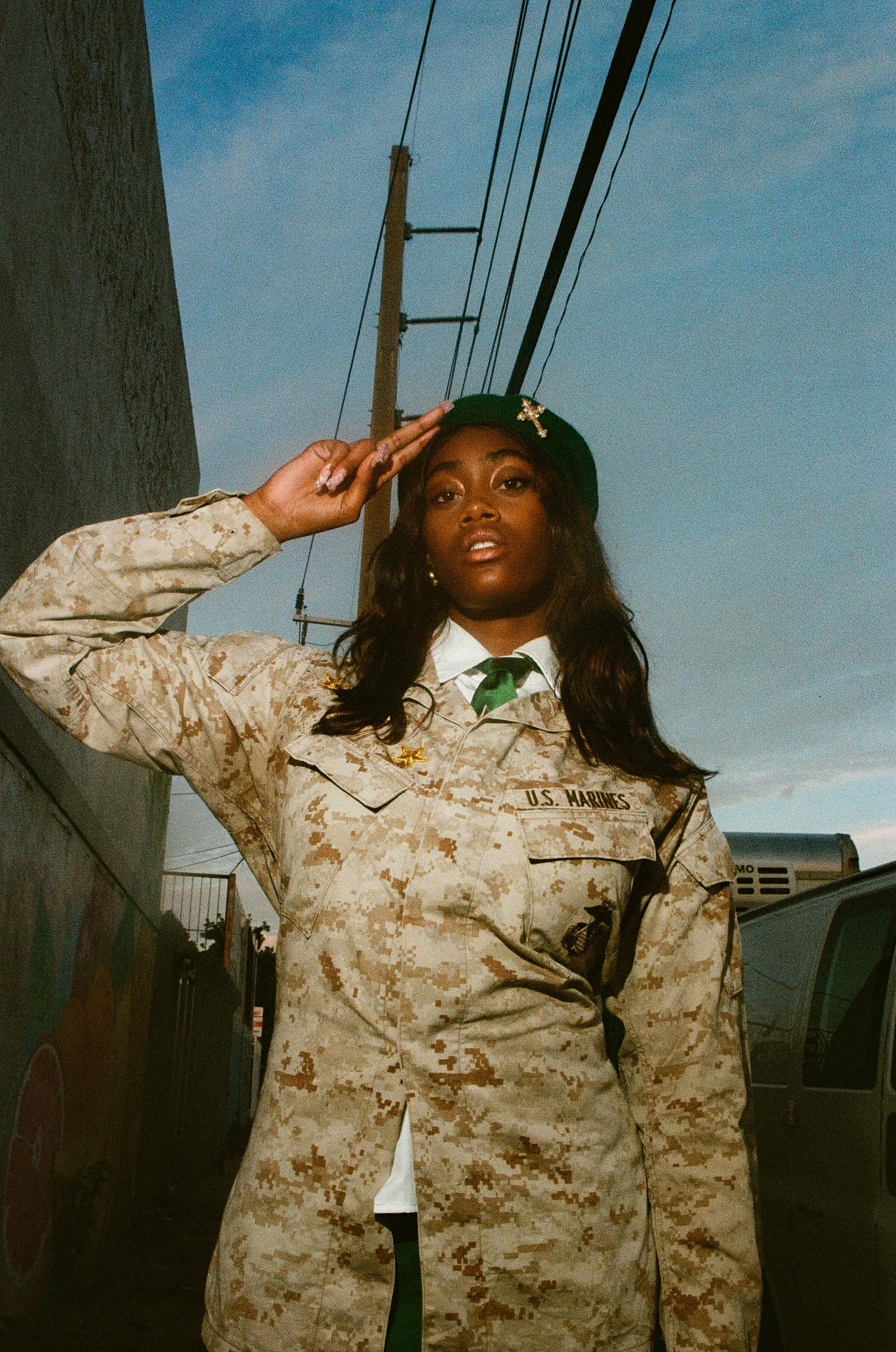 A woman in a U.S. Marines desert camouflage uniform salutes outdoors during dusk, with power lines and a building in the background.