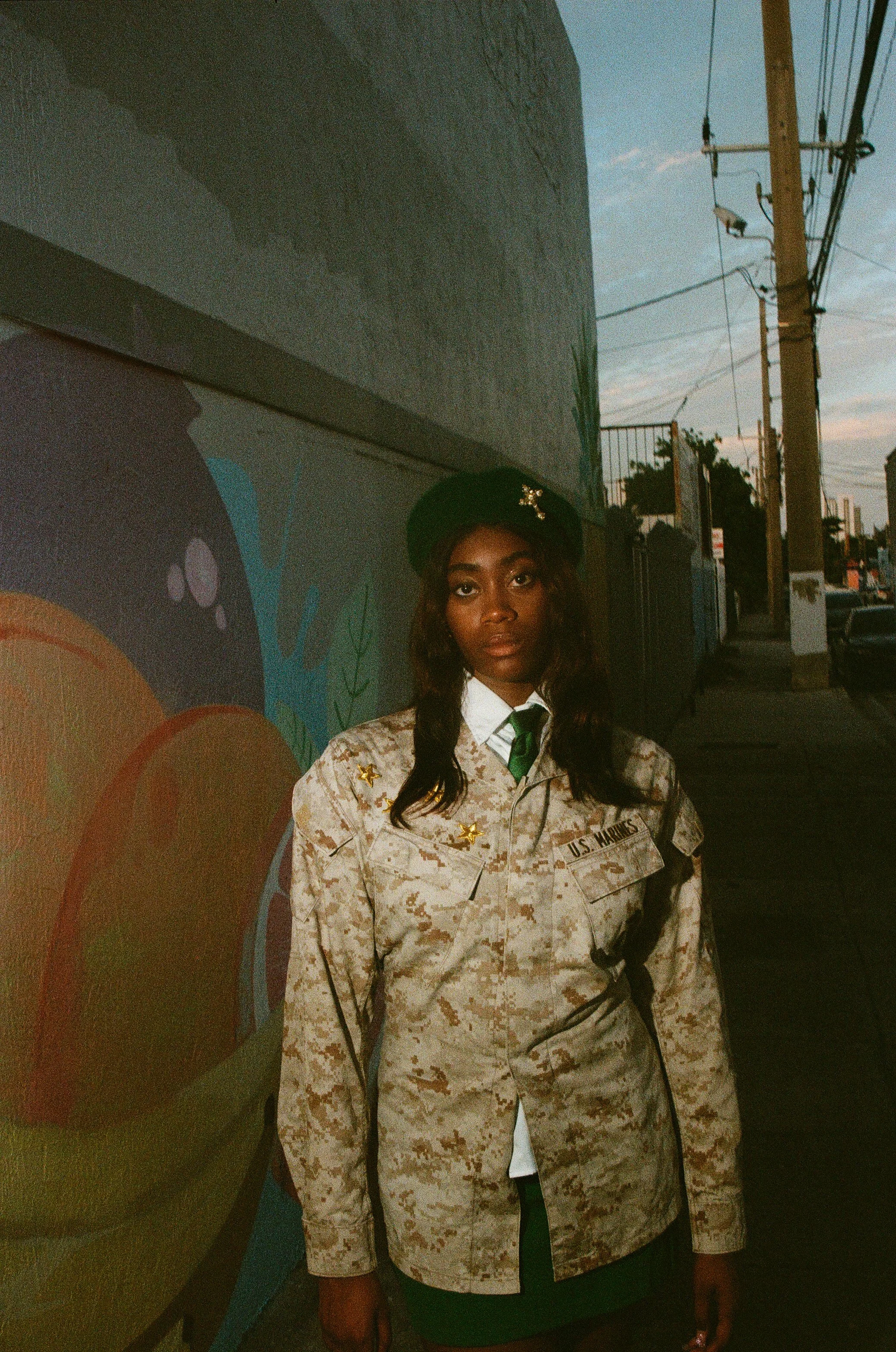 A woman in camouflage military uniform, wearing a black beret with a star emblem, standing on a sidewalk at dusk against a colorful mural wall with urban street and power lines in the background.