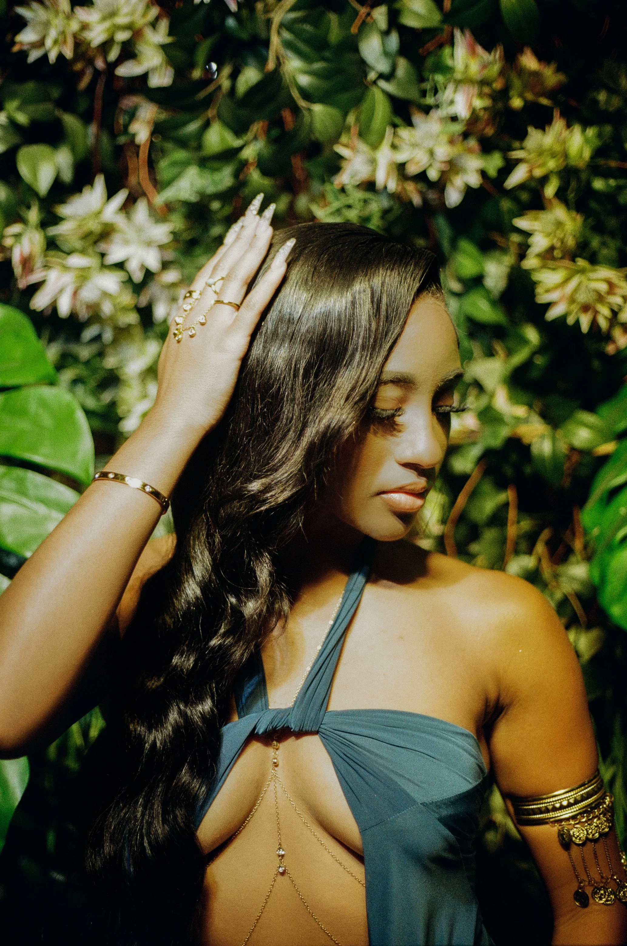 A woman with long wavy black hair wearing a dark teal dress with a unique strap design, and gold jewelry, standing against a lush, green leafy background.