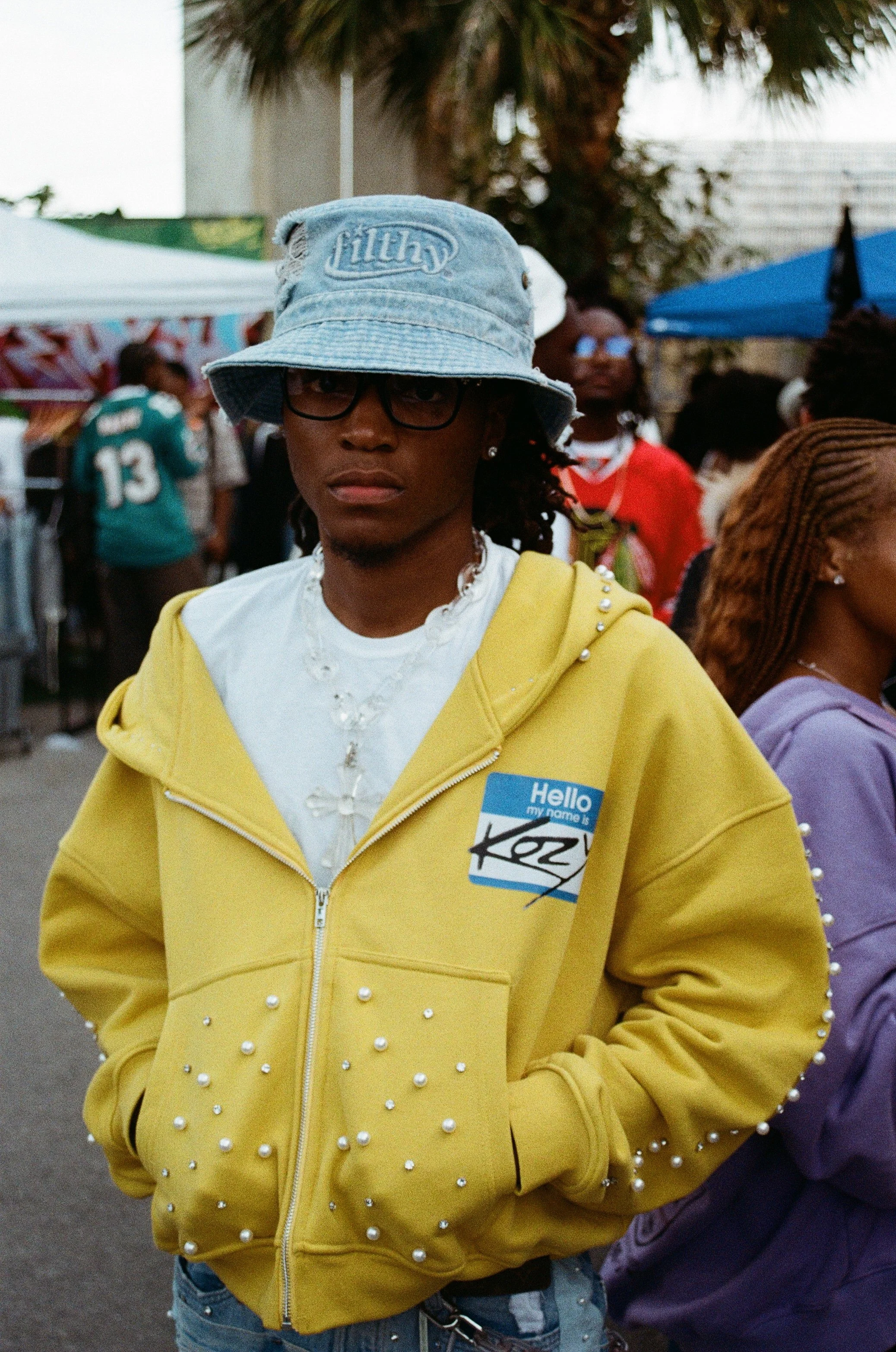 A young man wearing a light blue bucket hat, black glasses, a yellow hoodie with pearl embellishments, and a white t-shirt with a cross necklace, standing at an outdoor event with several people and tents in the background.