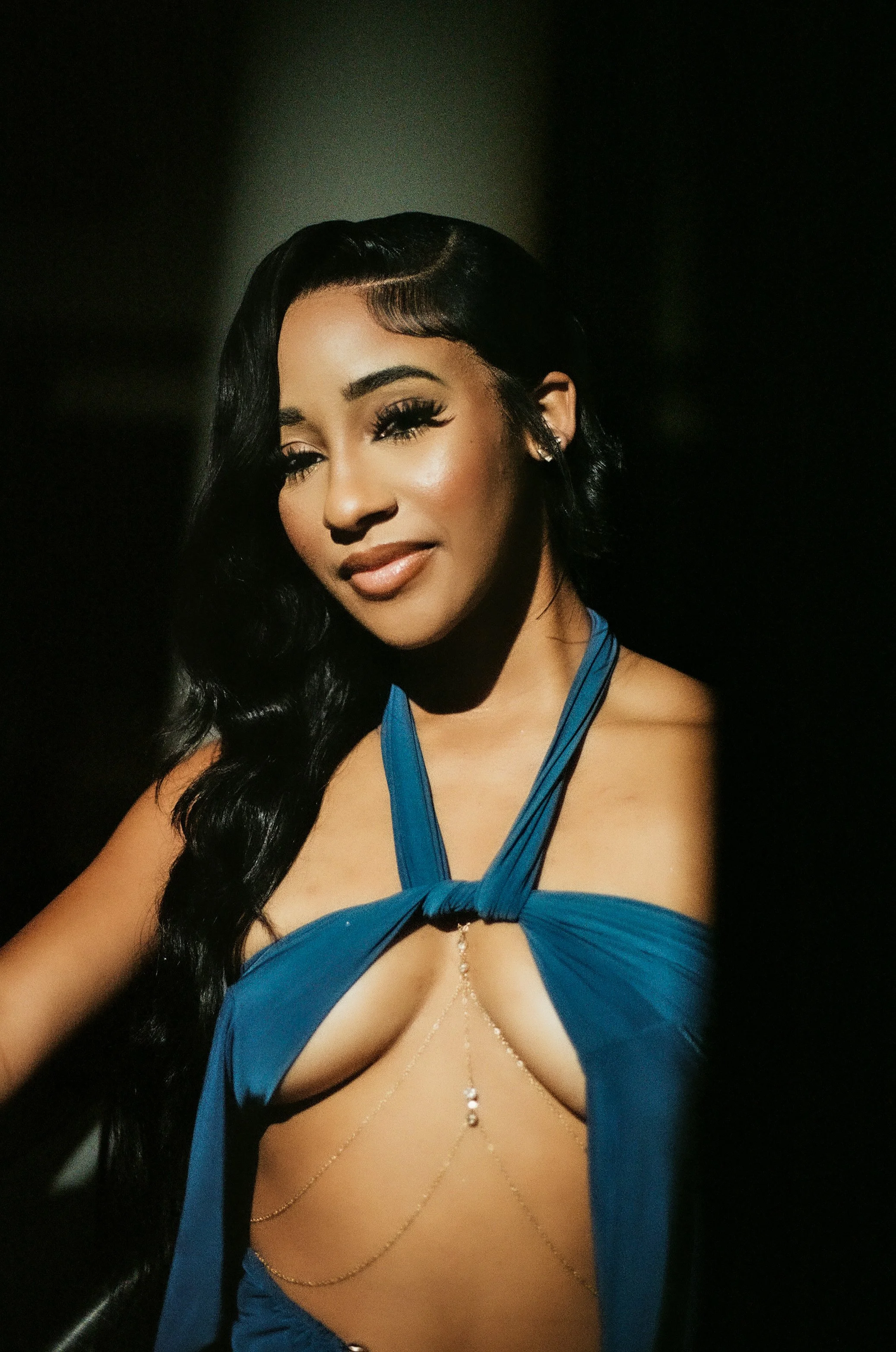 A woman with long black hair, wearing makeup, and a blue dress with chains, smiling at the camera against a dark background.