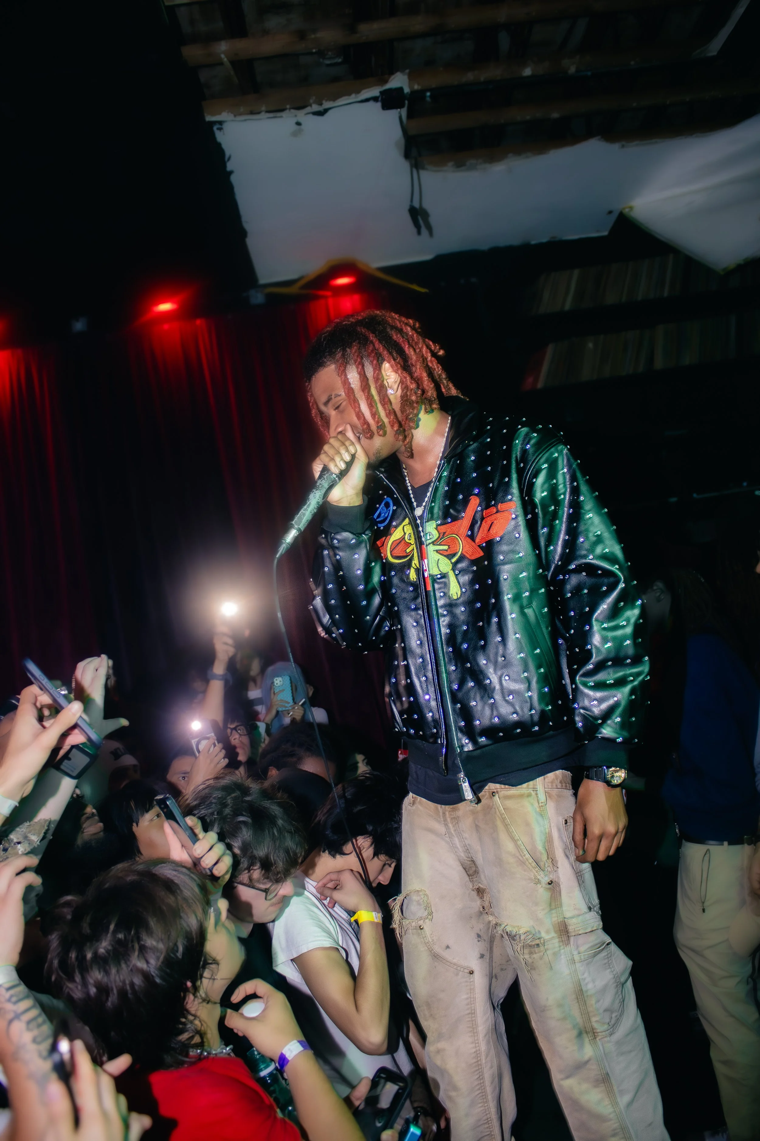 Performer with red dreadlocks singing into a microphone on stage at a concert, surrounded by a crowd taking photos and videos.