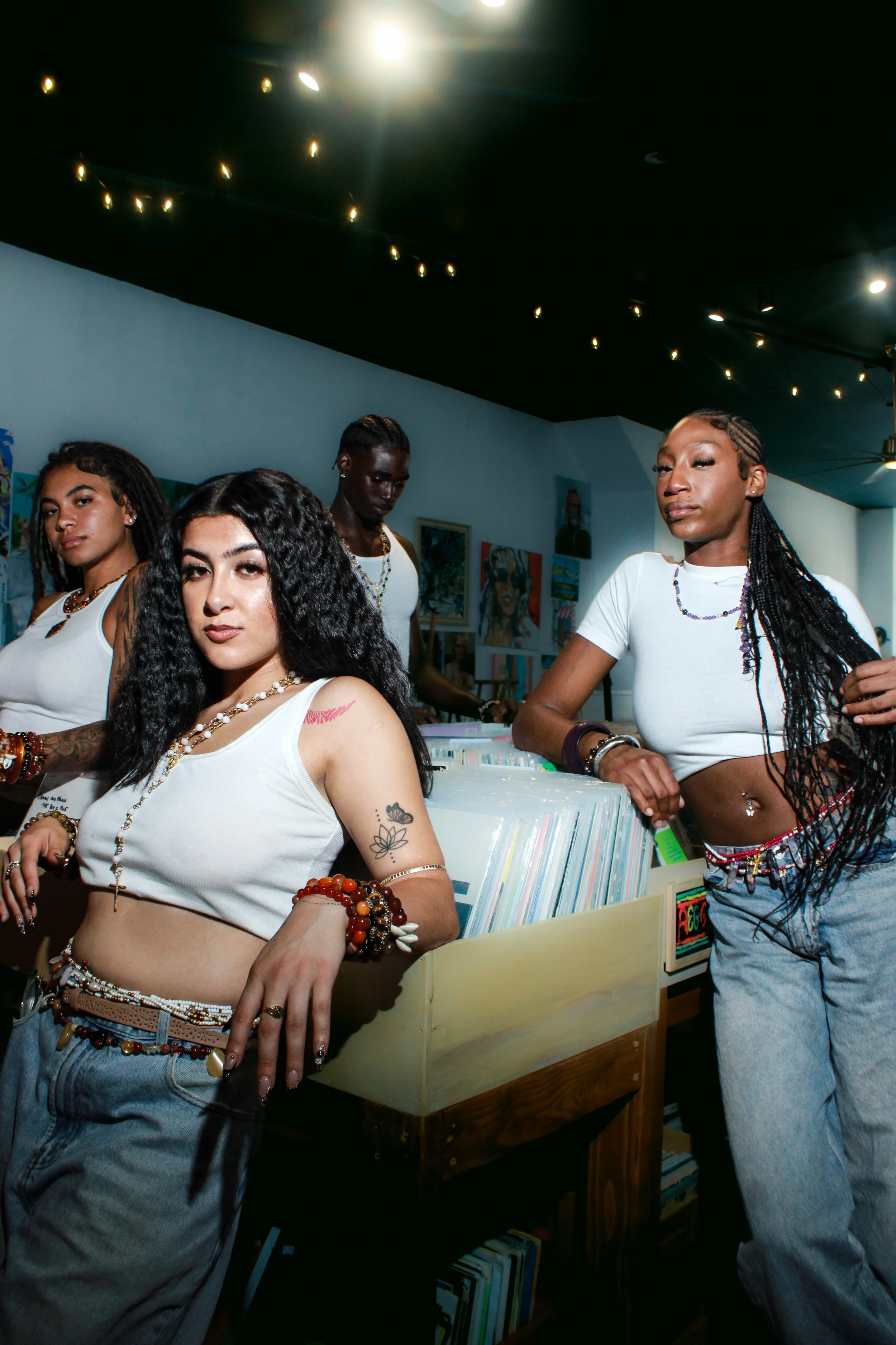 Group of five young women at a record store, some leaning on records and others standing, with artwork on the walls and string lights on the ceiling.