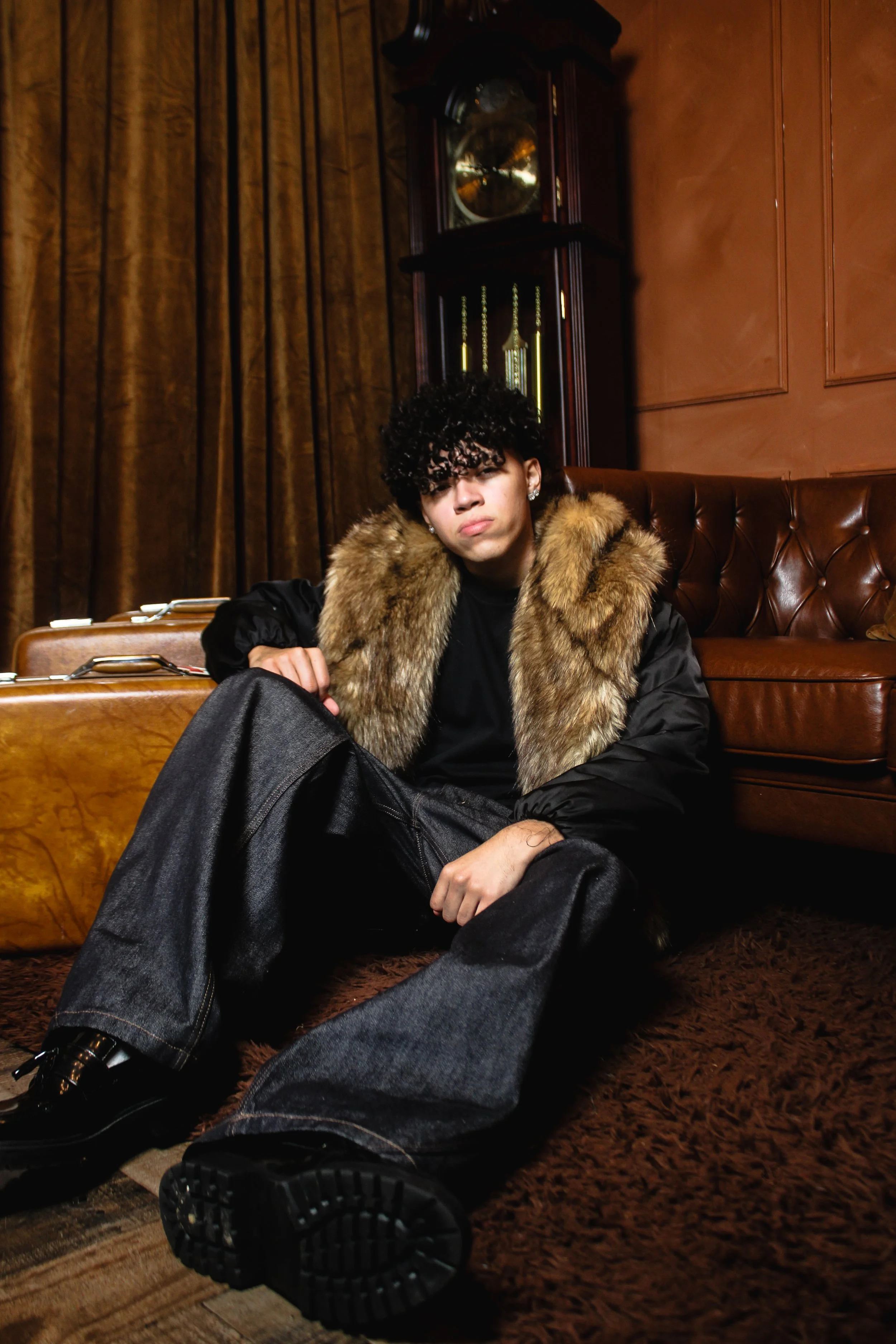 A young person with curly black hair and earrings sitting on the floor in a room with brown wood paneling and a tall grandfather clock. They are wearing a black shirt, dark pants, shiny black shoes, and a jacket with a fur collar.
