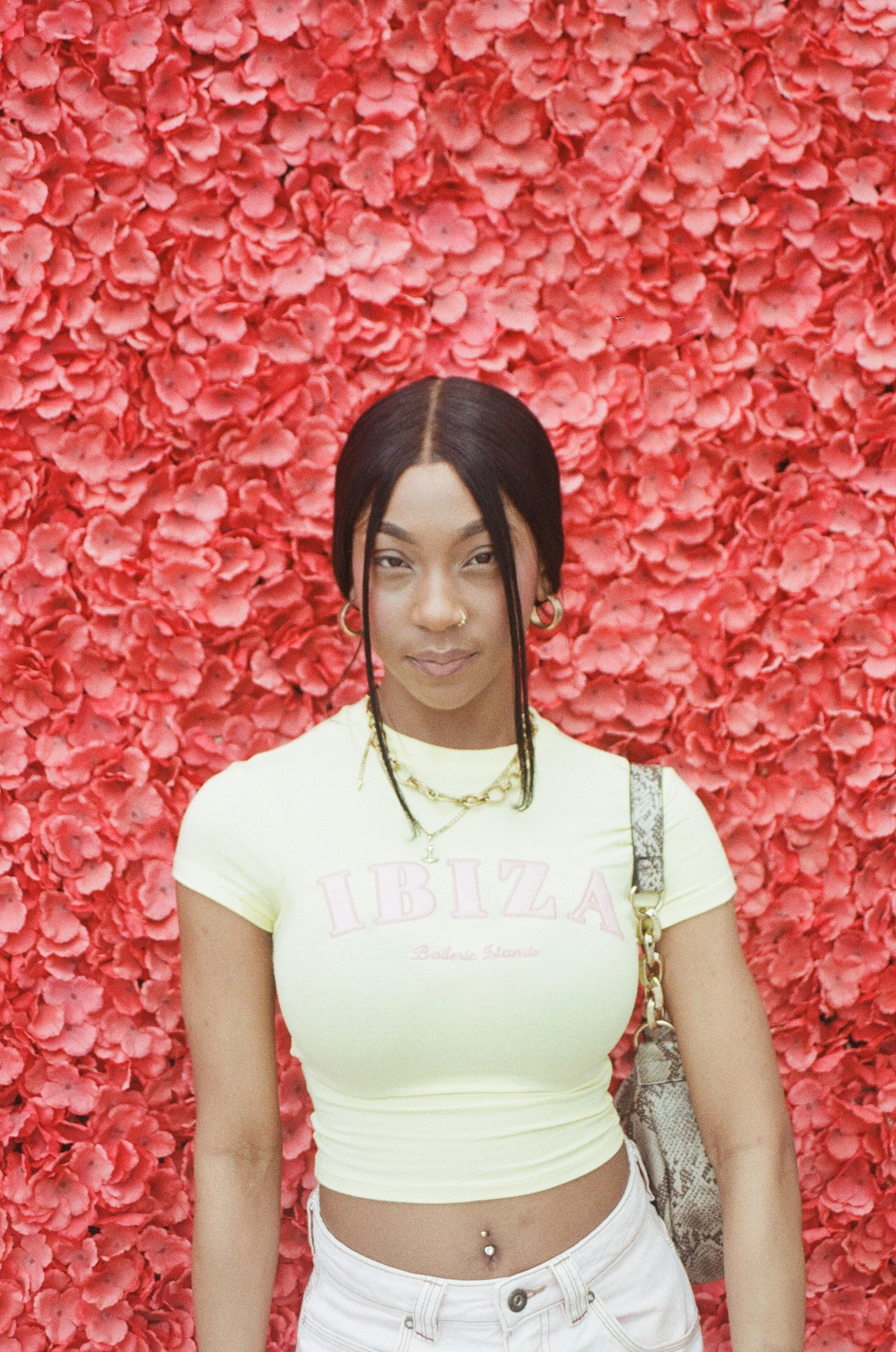 A woman with dark hair and gold jewelry posing in front of a wall of pink flowers, wearing a yellow "Ibiza" crop top and white pants.