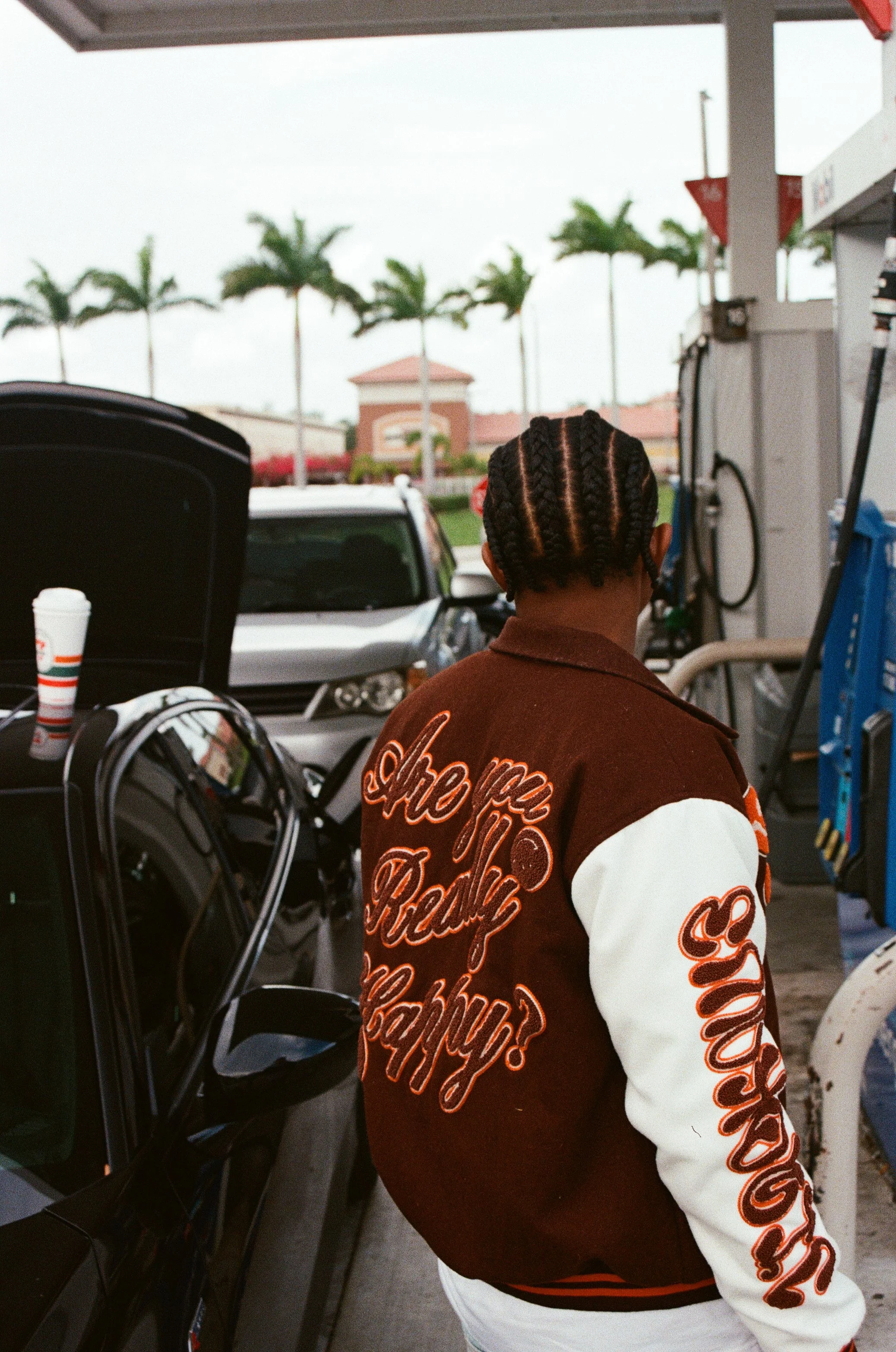 Back of a person with braided hairstyle at a gas station, wearing a brown and white jacket with orange lettering, filling up a black car with a soda cup on the open hood. Palm trees and a shopping center are visible in the background.