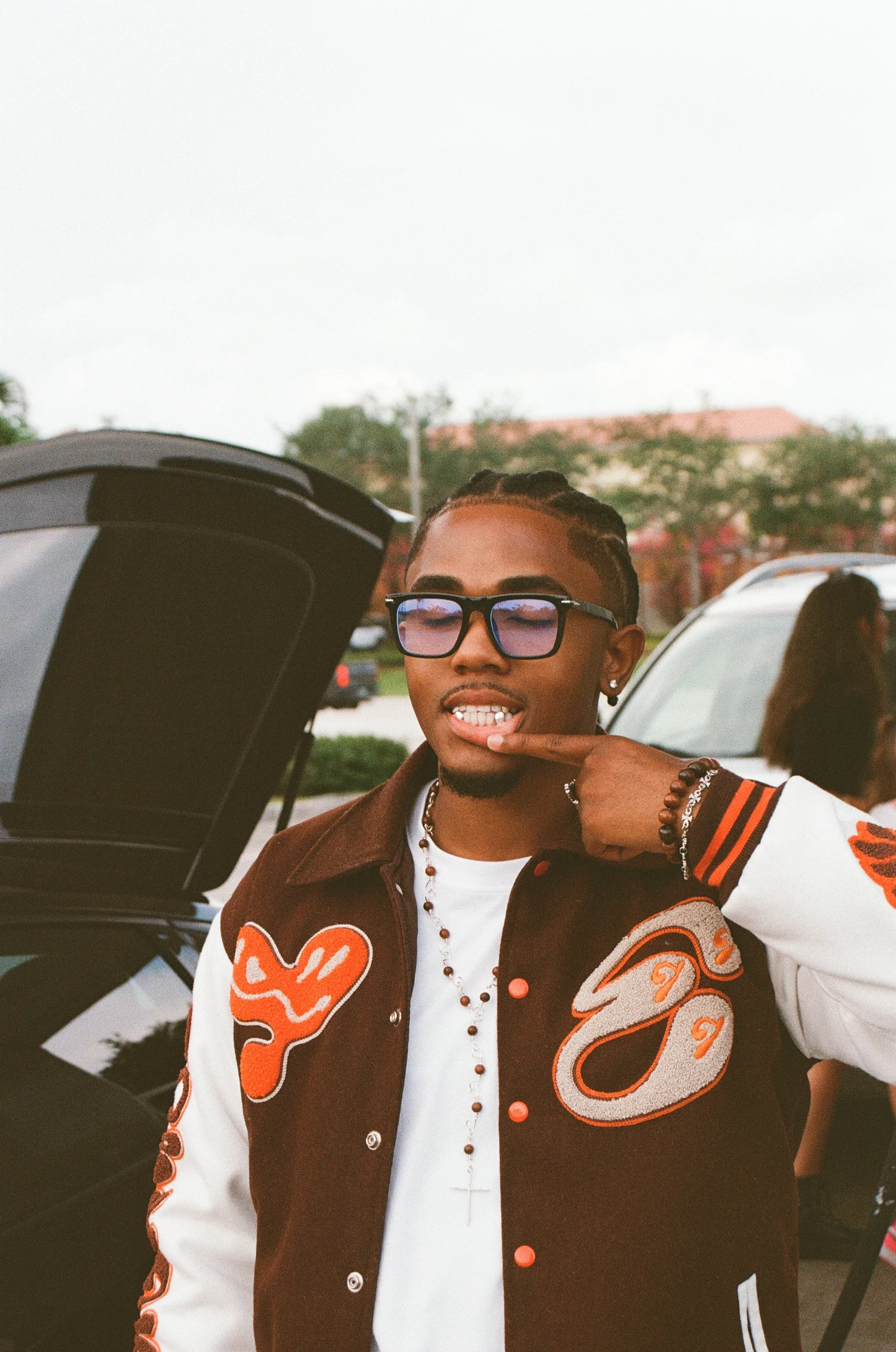 A young man with braids in a varsity jacket with orange and white patches, wearing black glasses with reflective lenses, silver earrings, layered necklaces, showing his grill while pointing to his mouth, standing outdoors in a parking lot.