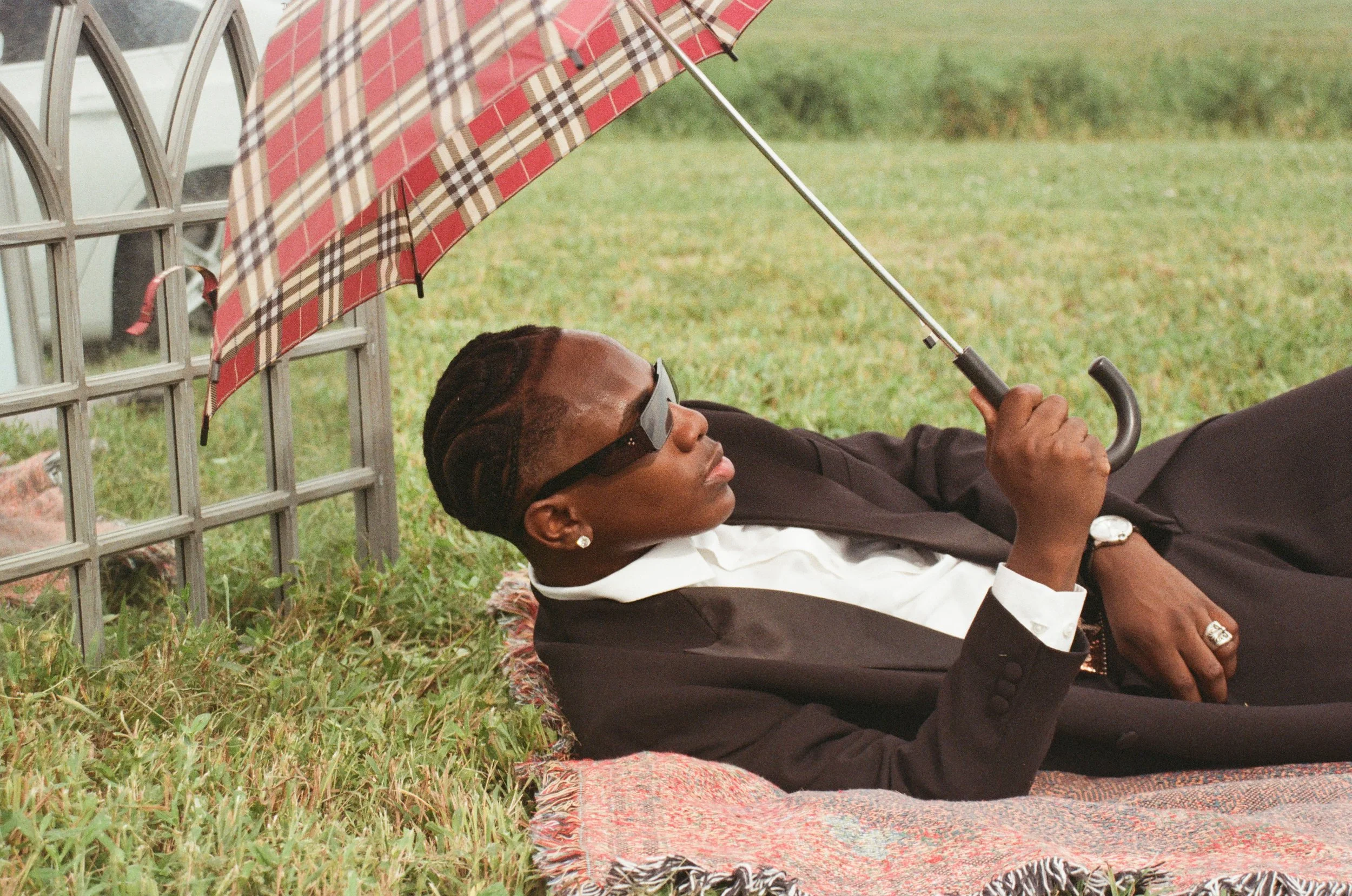 A man in a suit relaxing on a colorful rug outdoors, lying on his back under a plaid umbrella, wearing sunglasses and a watch, with a ring on his finger, holding the umbrella's handle, with grass and a vehicle in the background.