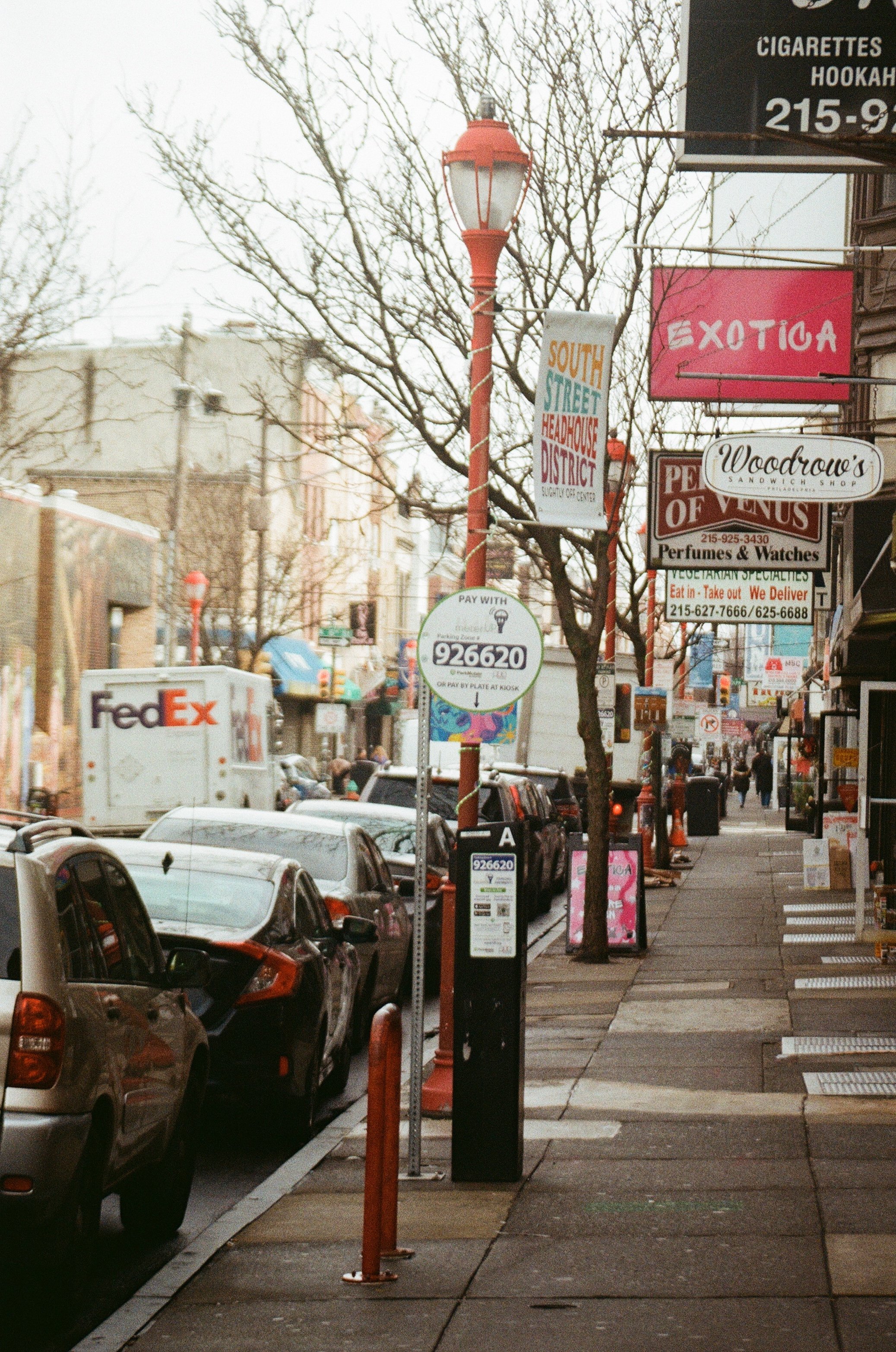 City sidewalk with parked cars, street lamps, and various store signs, including a sign for cigarettes and hookah, a sign for exotic items, and a pizza shop, in an urban area.