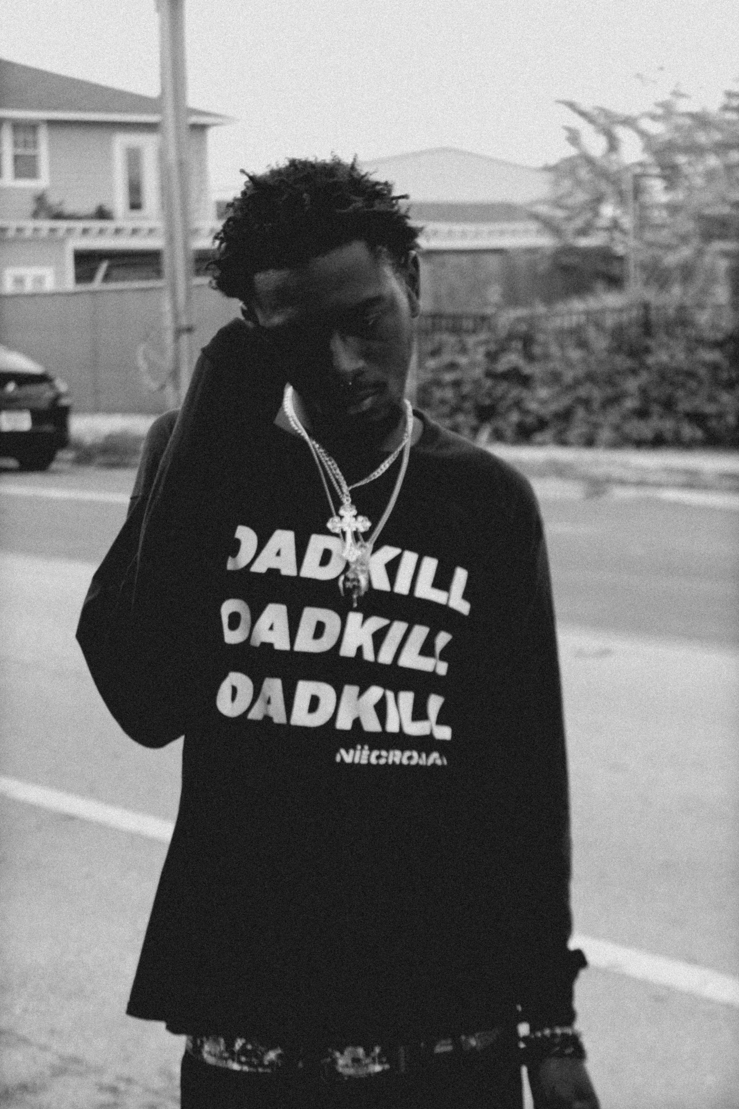 A young man with dreadlocks wearing a black hoodie with the word 'DADKILL' repeated multiple times, standing outdoors on a street, wearing chains, in black and white.