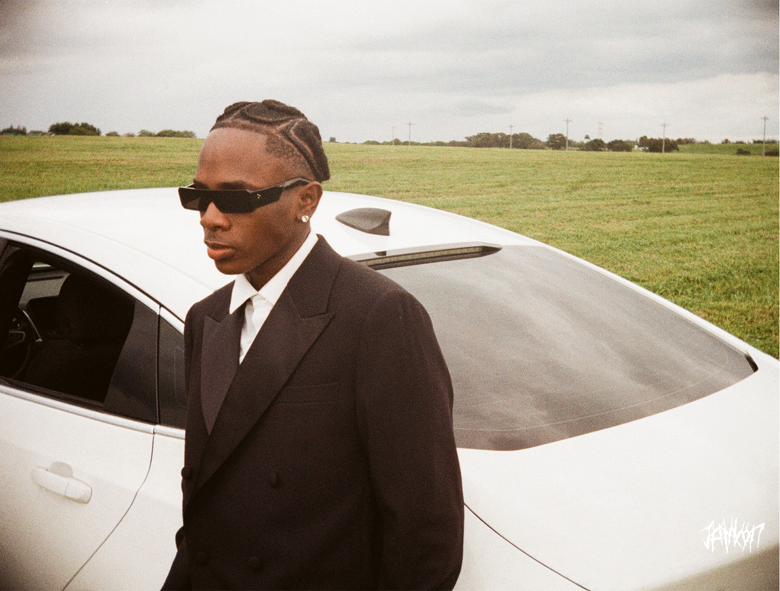 A man in a black suit and sunglasses standing beside a white sports car in an open grassy field on a cloudy day.