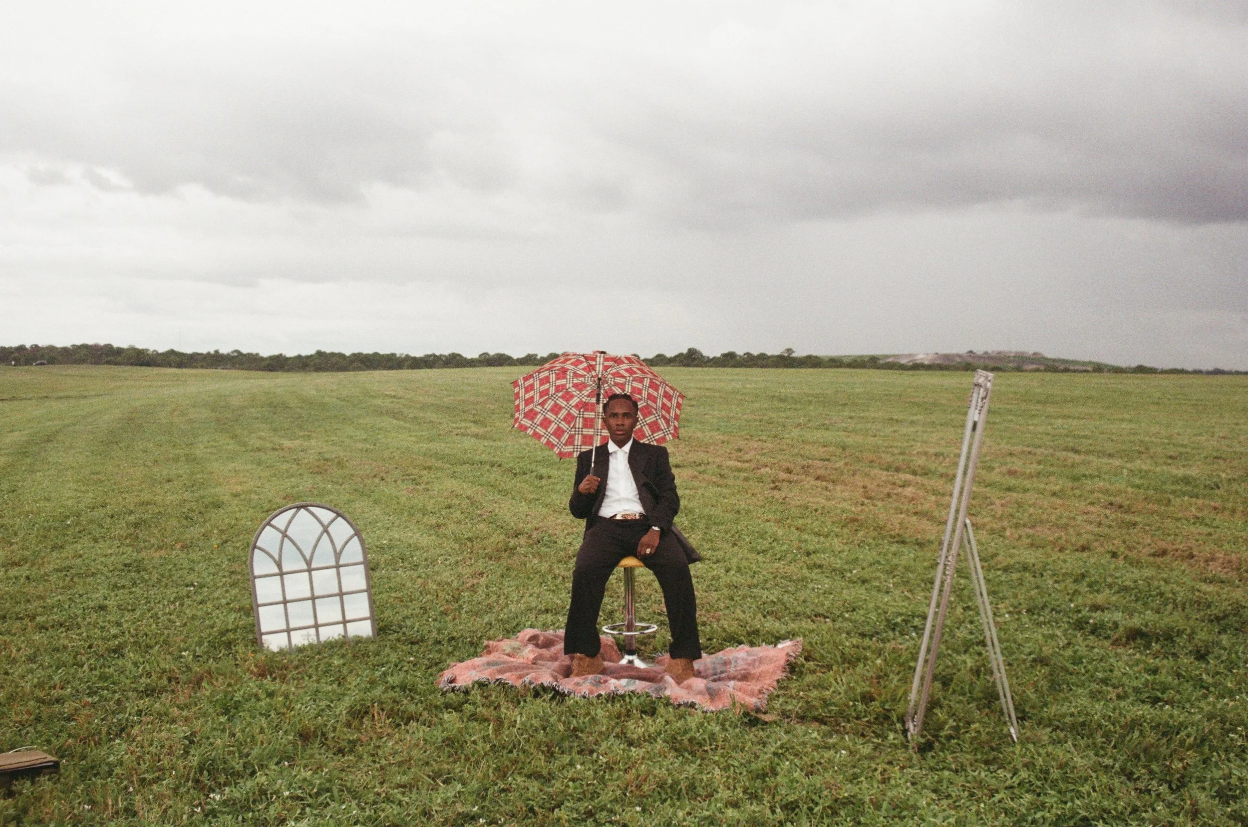 A man in a suit sitting on a stool in a grassy field holding a red and white checkered umbrella. There is a decorative window frame on the ground to his left and a tall easel to his right. The sky is overcast.