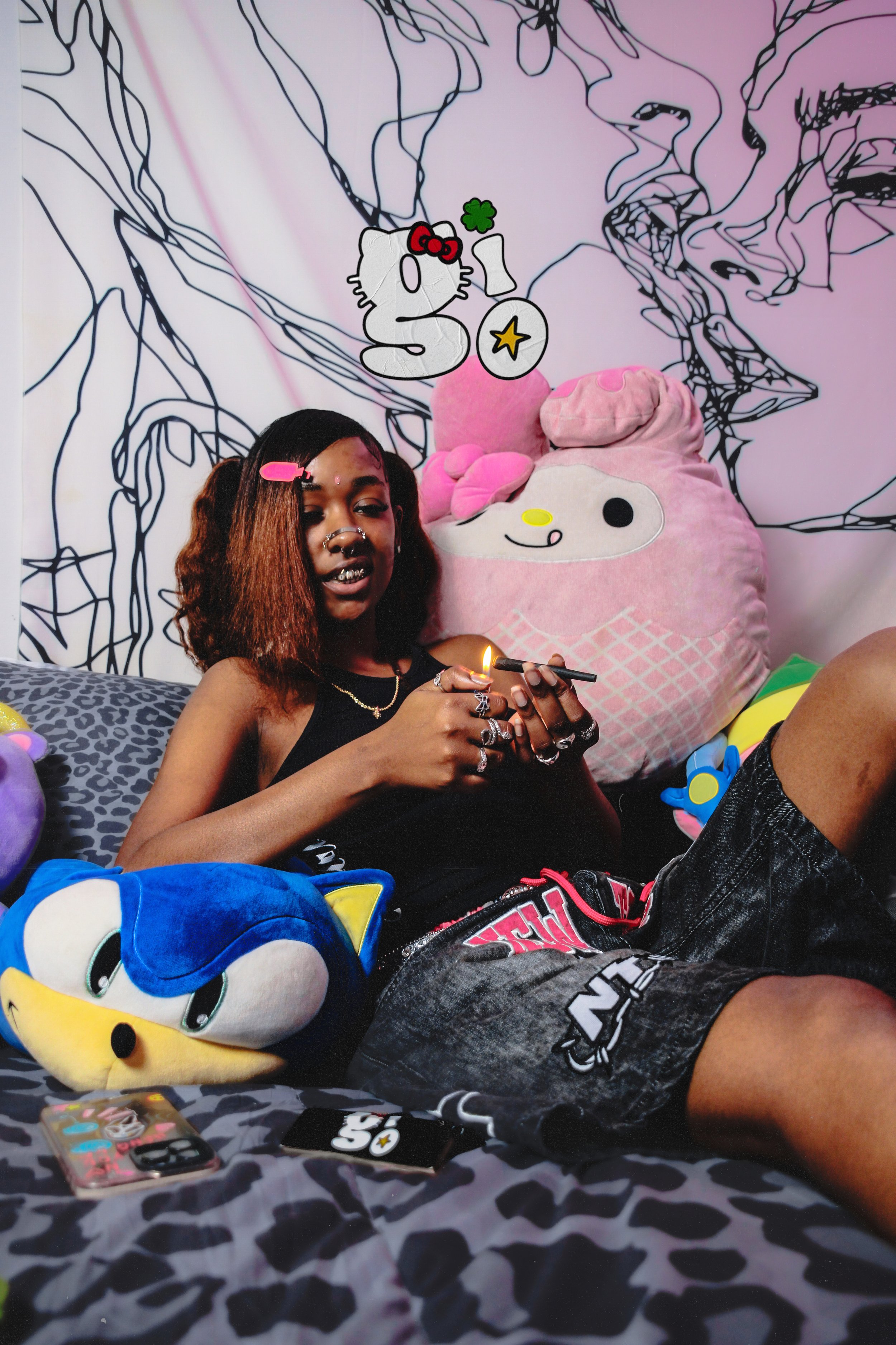 Young person with colorful hair and jewelry sitting on a bed with plush toys, including a Sonic the Hedgehog stuffed animal, in a room decorated with black line art on a white wall and Hello Kitty stickers flying around.