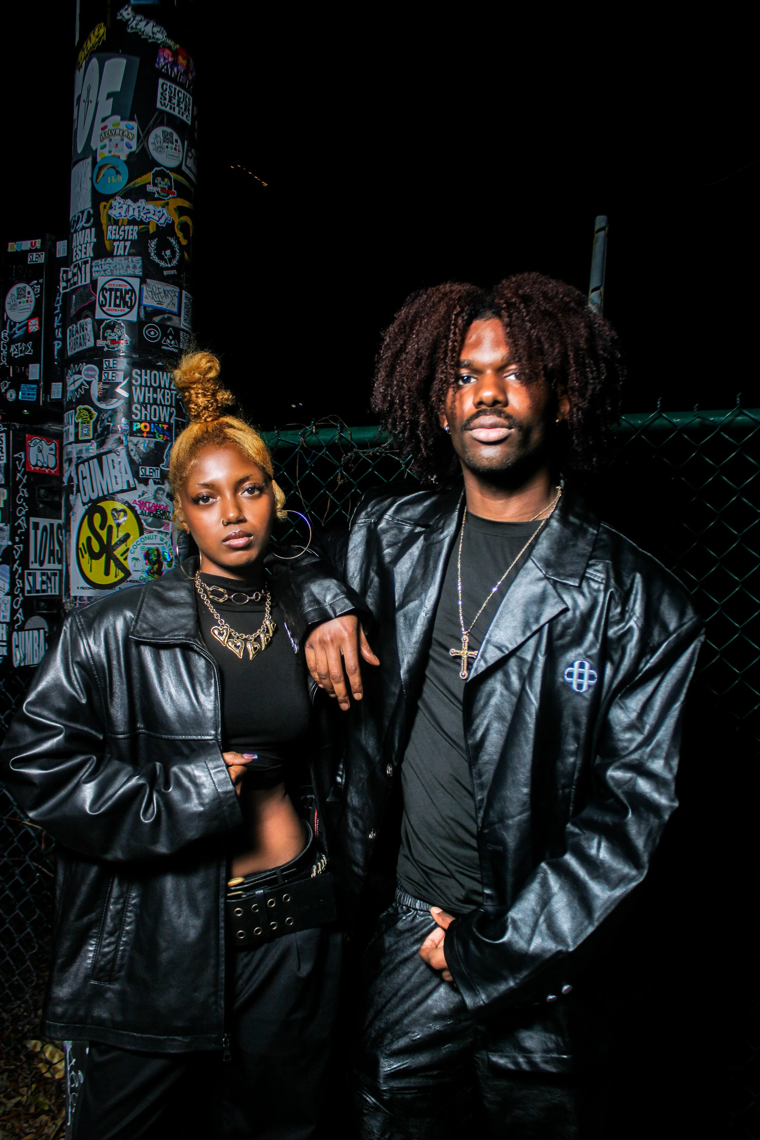 Two young adults, a woman and a man, standing in front of a chain-link fence at night, wearing black leather jackets and jewelry, with stickers on a pole and graffiti in the background.