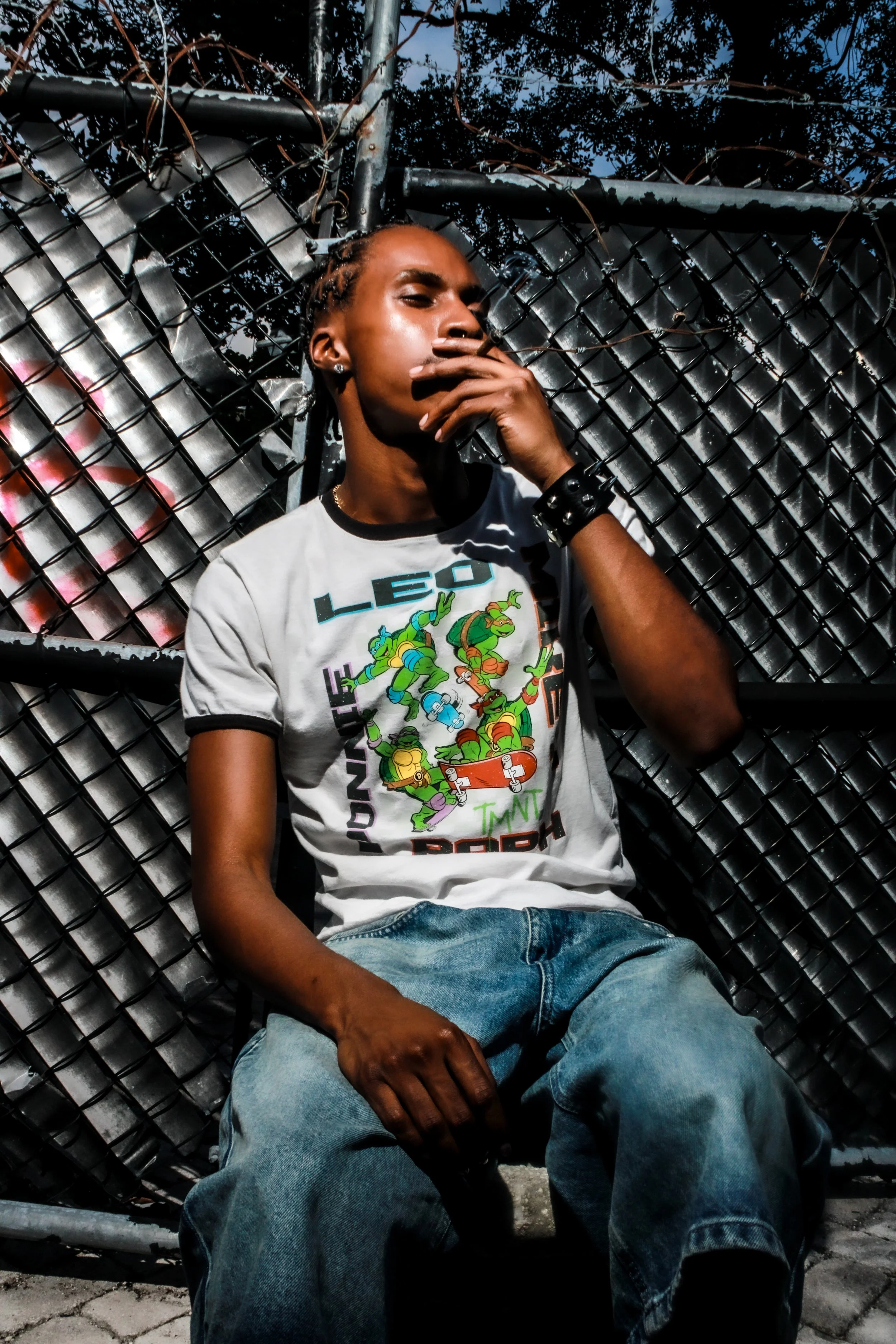 A young man sitting in front of a black metal fence with graffiti, wearing a Teenage Mutant Ninja Turtles t-shirt, jeans, and a black wristband, striking a contemplative pose with his hand near his face.