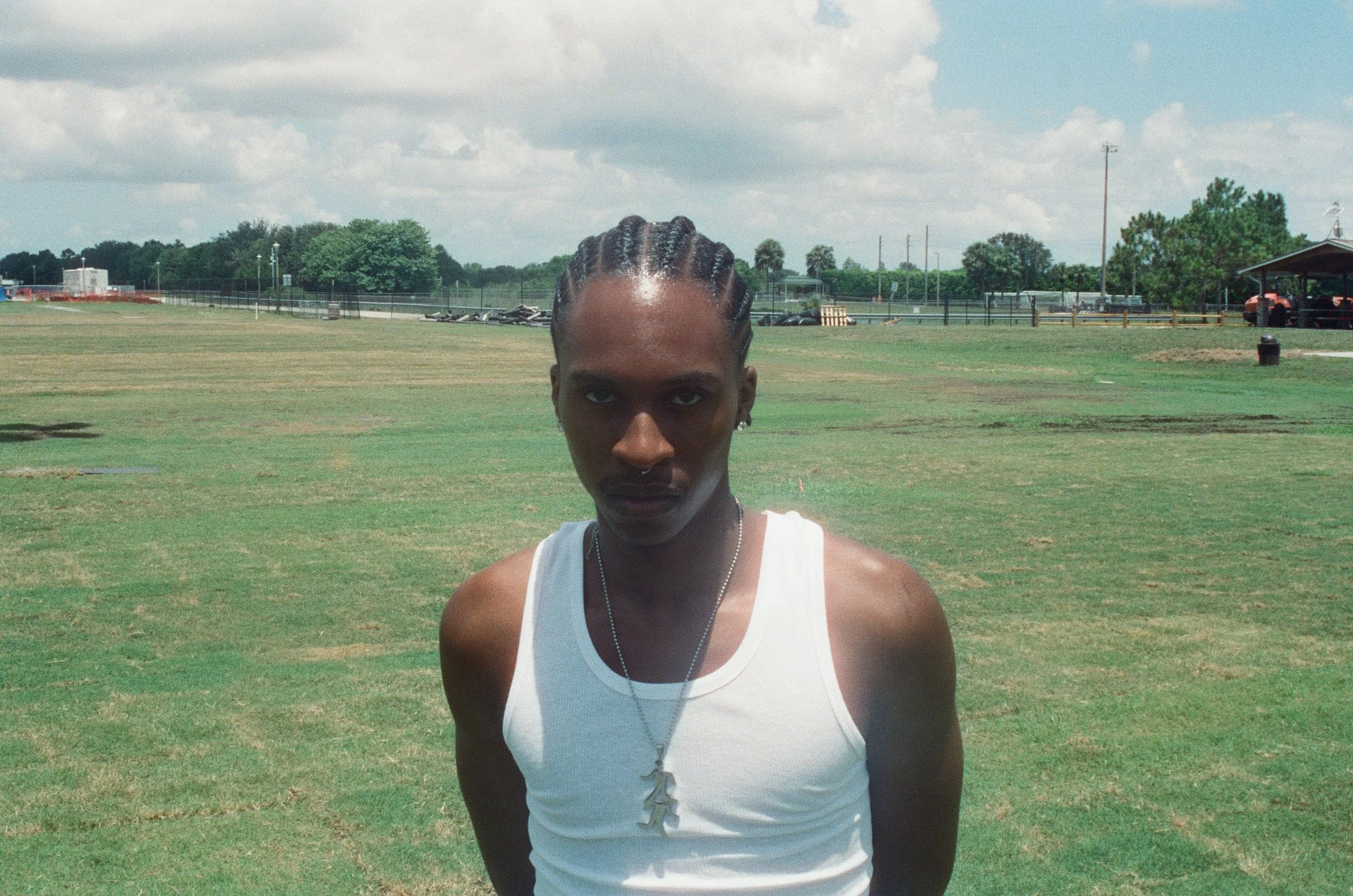 A young man with braided hair and dark skin, wearing a white tank top and a necklace with a cross pendant, standing outdoors on a grassy field under a partly cloudy sky.
